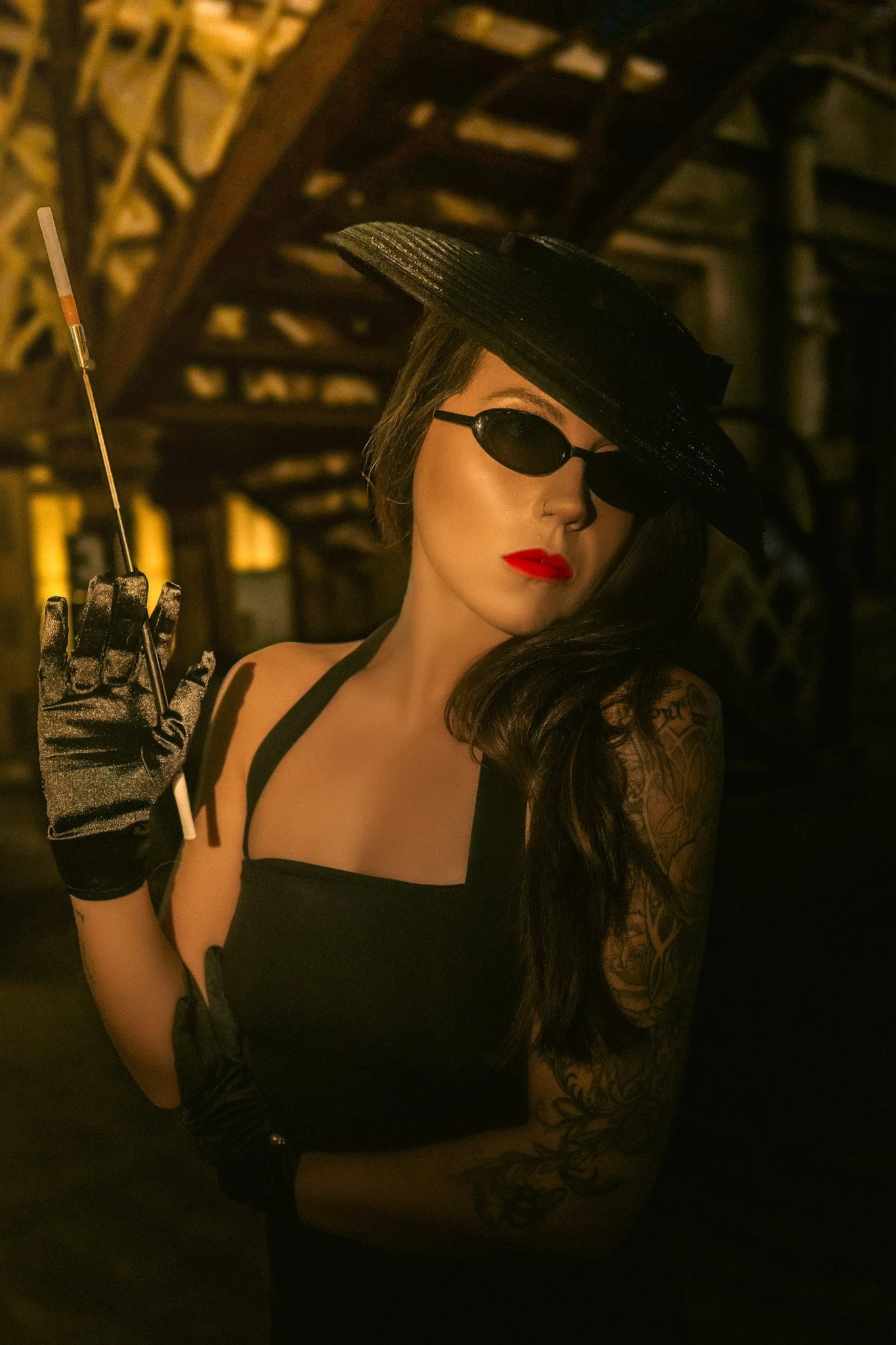 A woman with long dark hair, wearing a large black hat, oval sunglasses, red lipstick, a black sleeveless top, and black gloves, holding a thin cigarette holder, inside a dimly lit, industrial-style space.