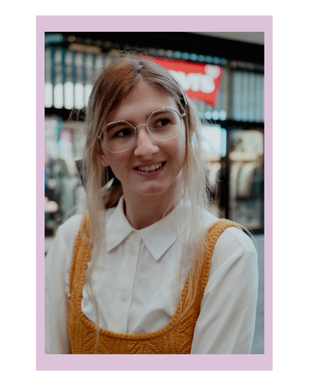 A young woman with light brown hair, glasses, and a white shirt with a yellow sweater vest, smiling at an indoor market or shopping area.