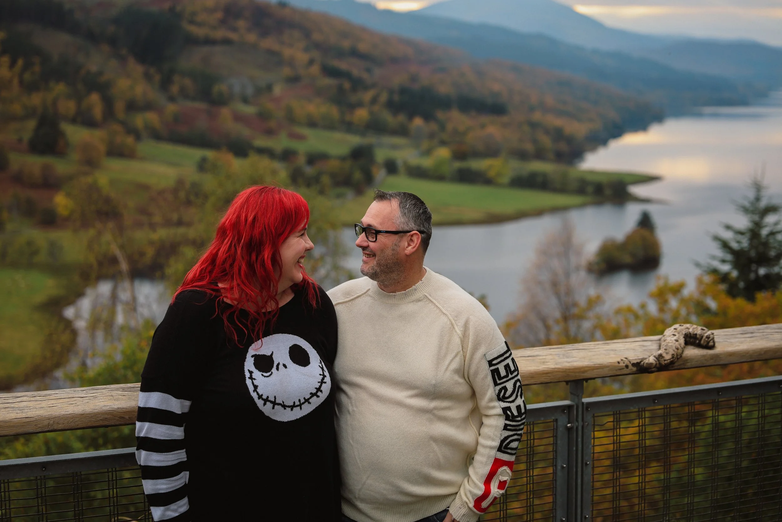 A smiling couple stands on a wooden observation deck overlooking a scenic landscape of a river, trees, and rolling hills during fall with colorful foliage.