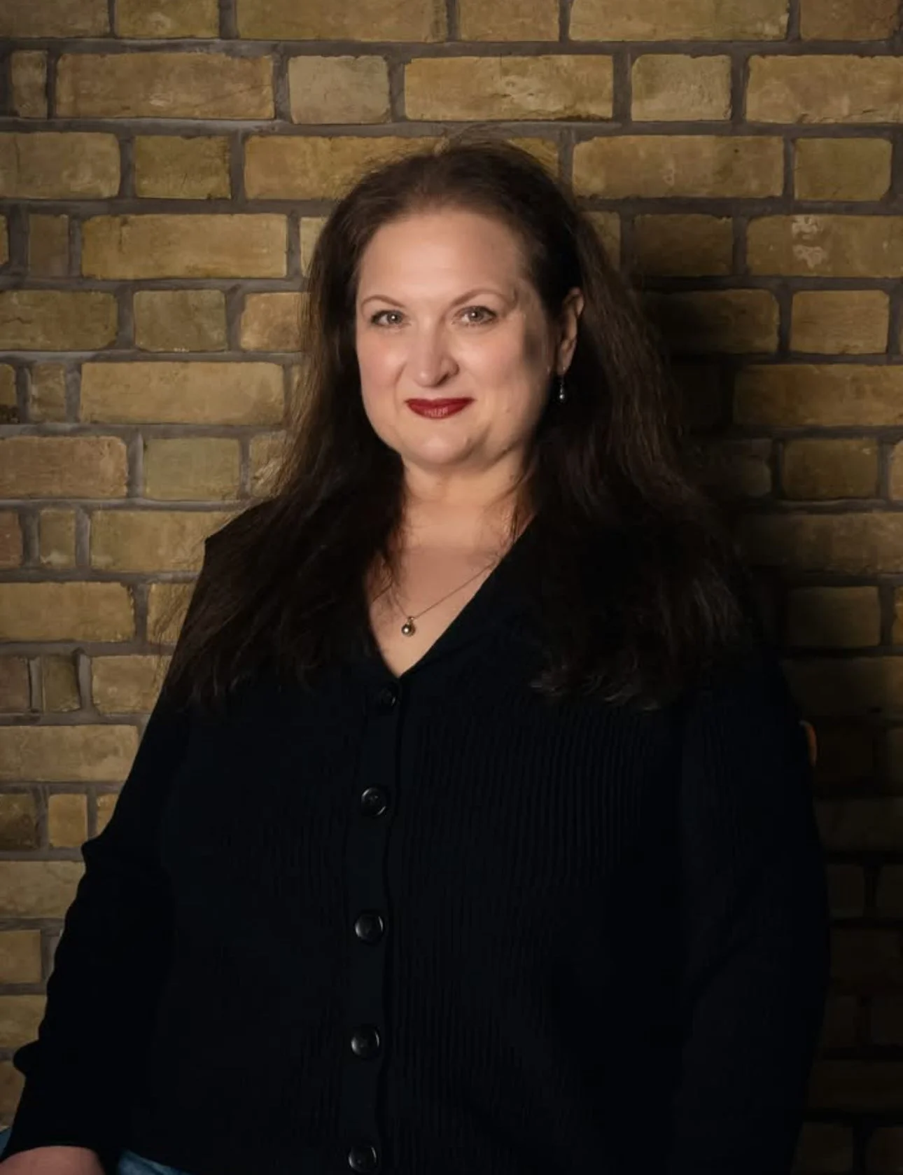 A woman with long dark hair wearing a black button-up sweater and jewelry standing against a brick wall.