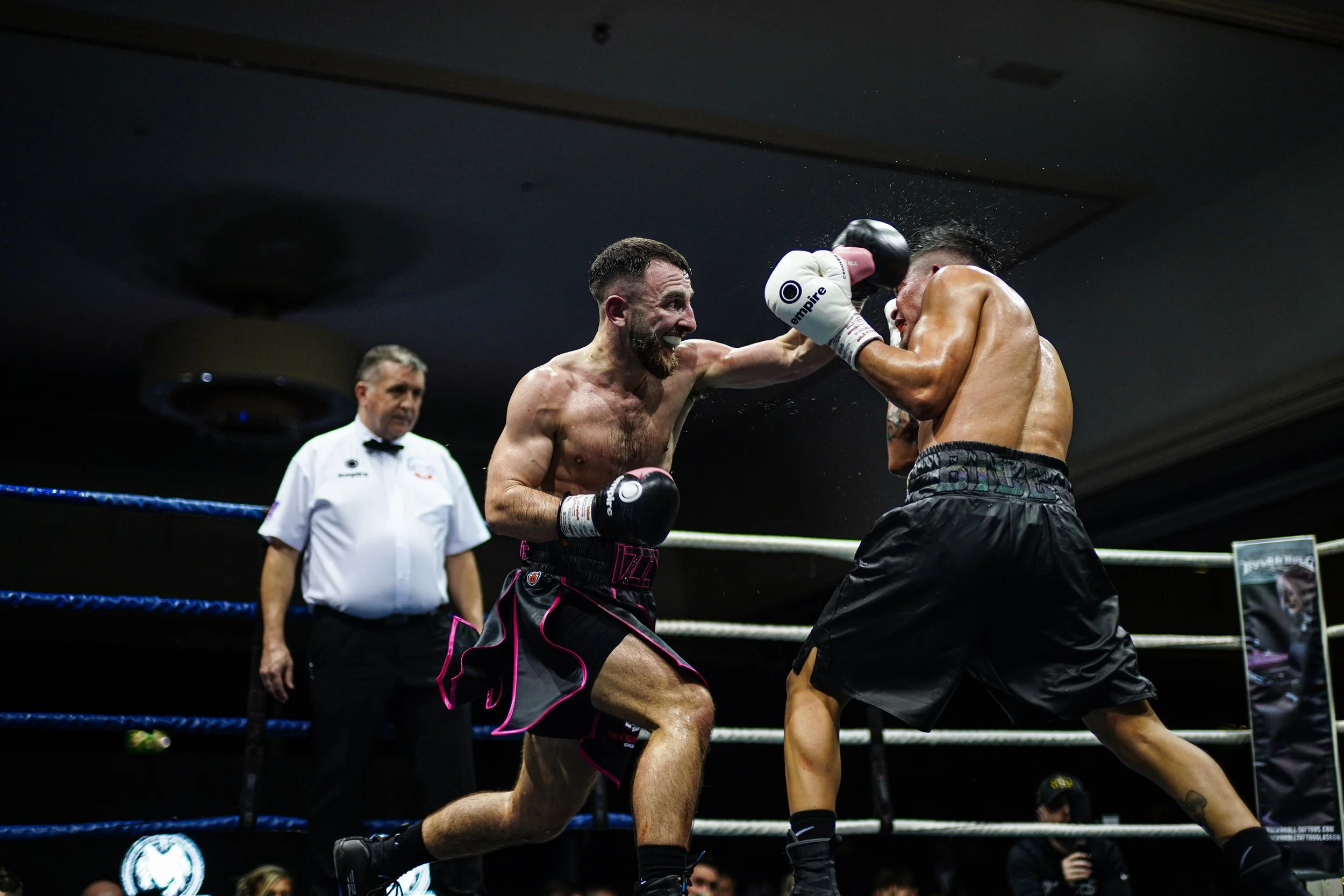 Two shirtless male boxers engaged in a fight in a boxing ring, with one delivering a punch to the other's face, while a referee observes in the background.