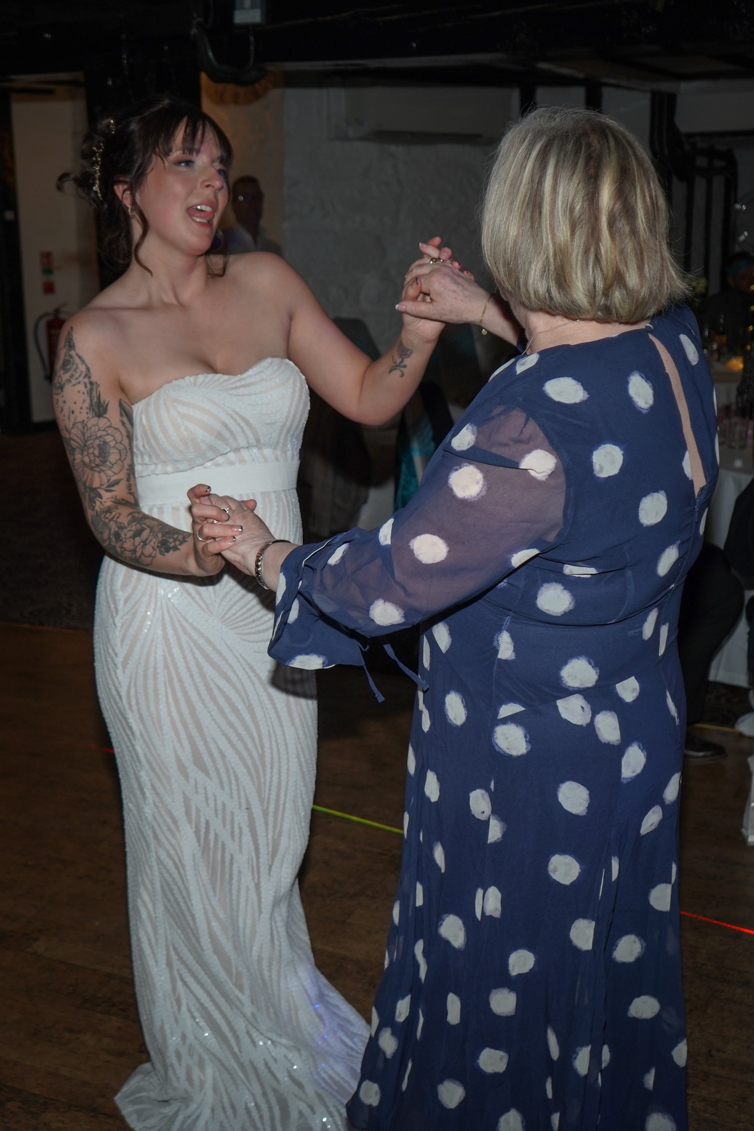 A young woman in a white strapless dress dances with an older woman in a blue dress with white polka dots at a celebration or wedding reception.