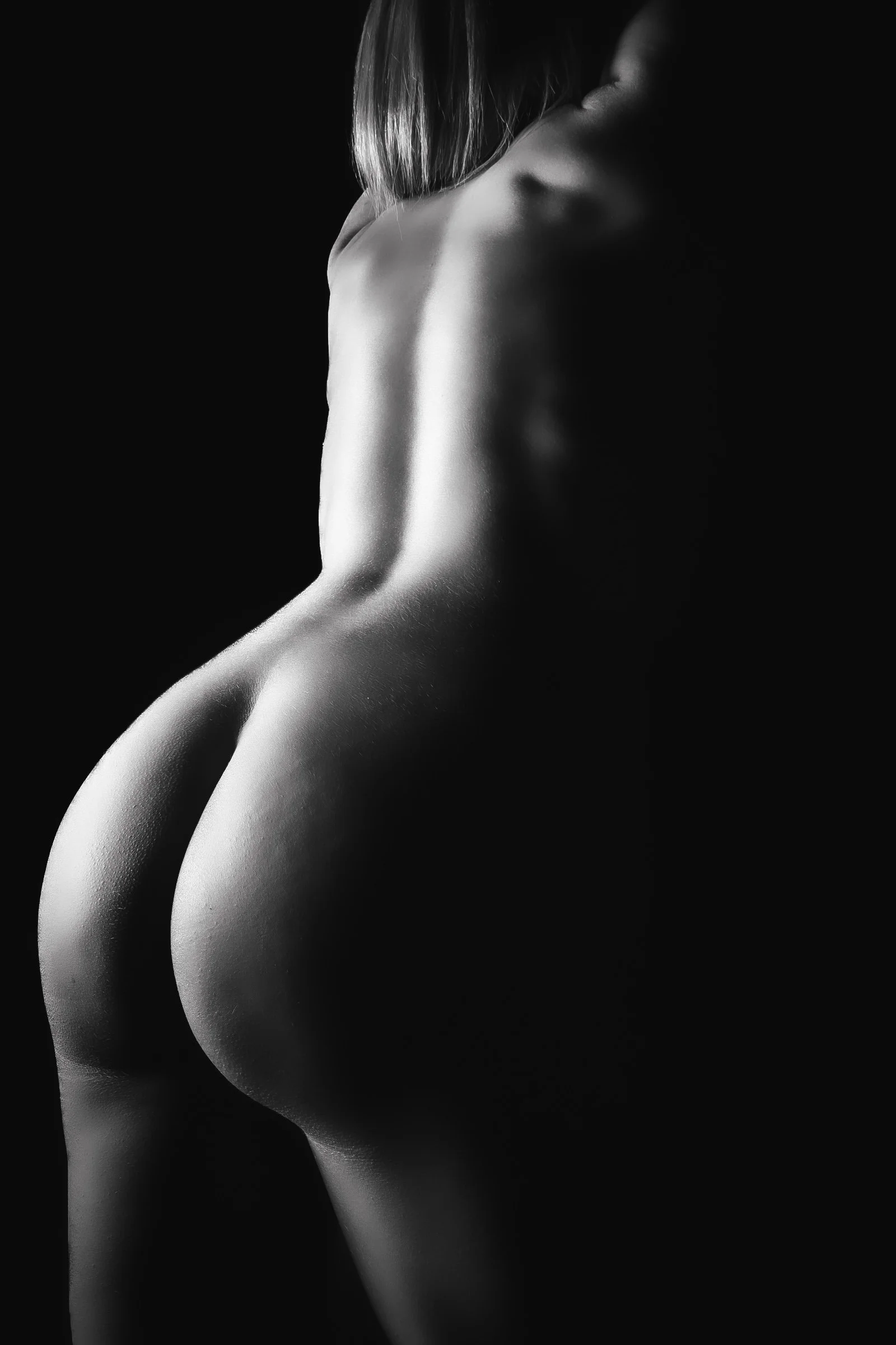 Black and white photograph of a person's back and buttocks, with the person facing away and partially turned to the side, illuminated by soft lighting against a dark background.