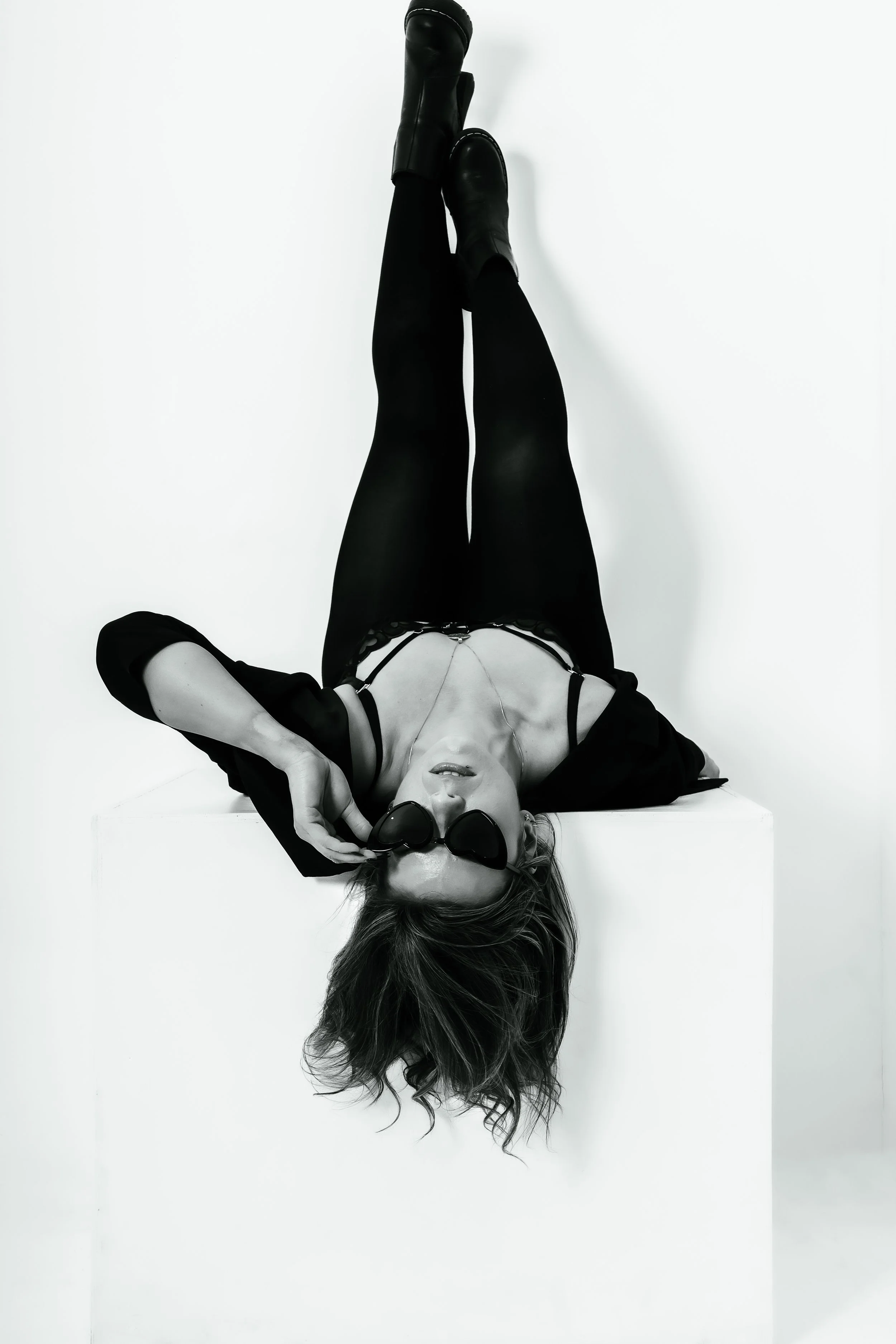 A woman lying upside down on a white block, holding sunglasses to her face, wearing black leather boots, black tights, and a black top against a white background.
