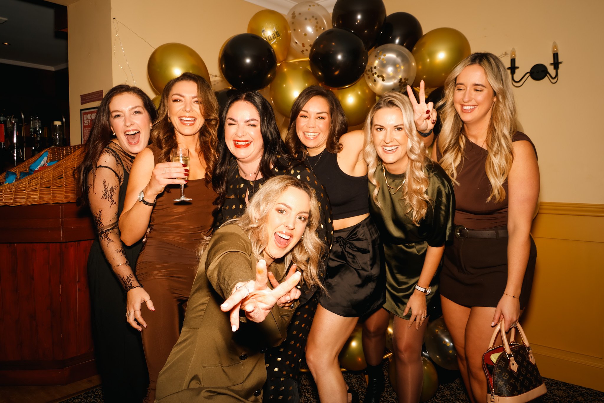Group of seven women celebrating at a party with gold and black balloons in the background, smiling and having fun.