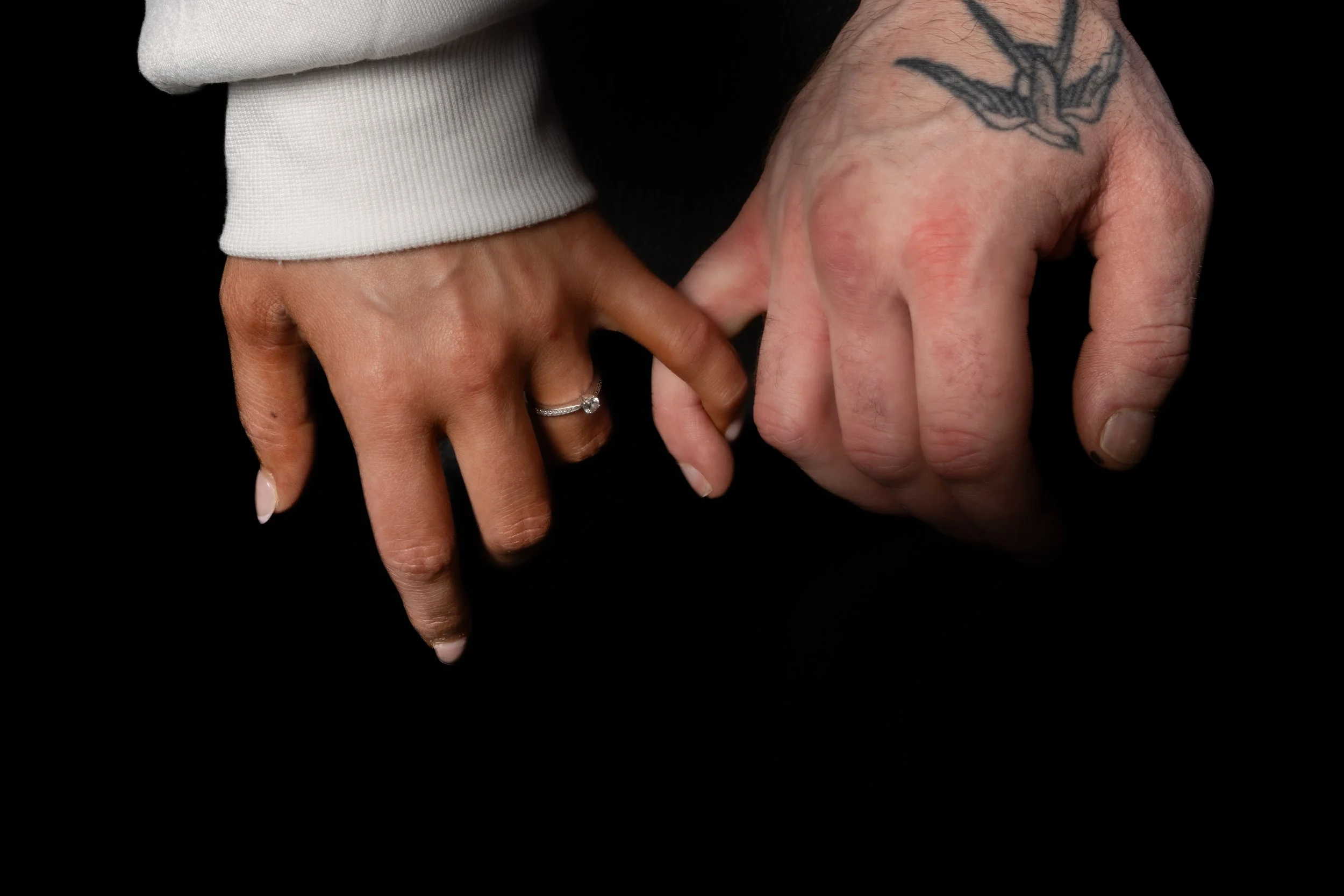 Two hands touching with fingers intertwined against a black background. One hand has a wedding ring and a tattoo on the wrist. The person on the left wears a white sleeve.