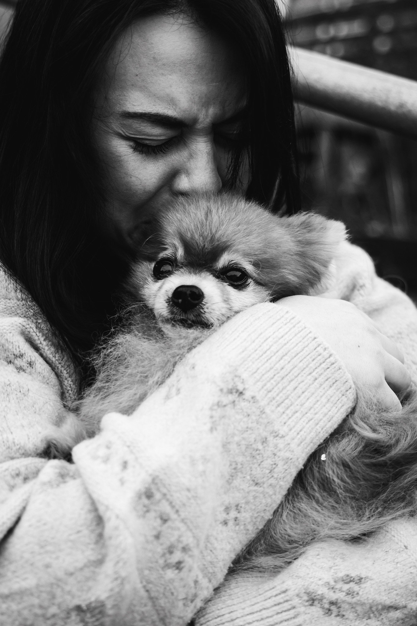A woman with dark hair hugging a small fluffy dog. She is kissing the top of the dog's head, and the dog has a calm expression. The photo is in black and white.