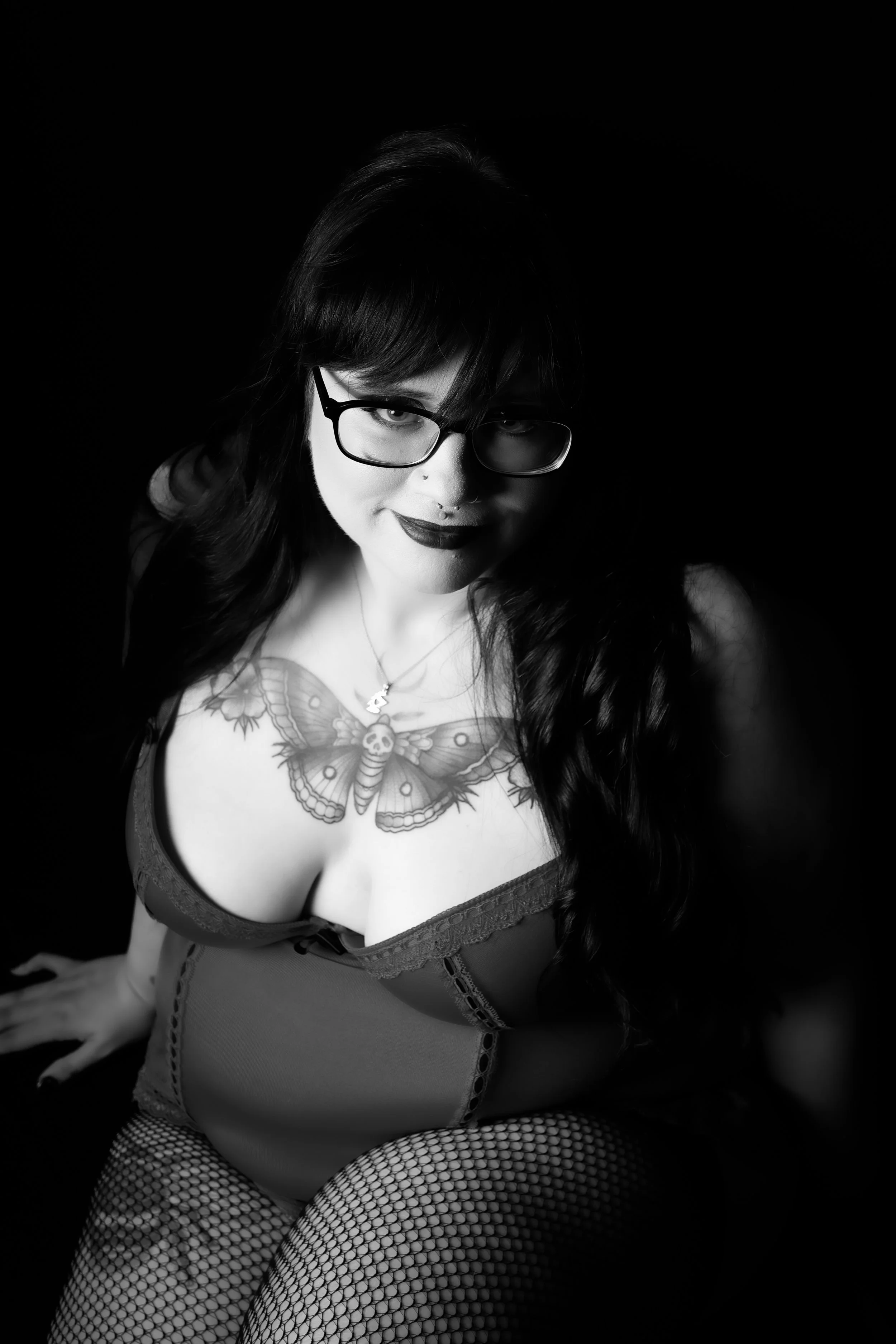 Black and white photo of a woman with dark hair, glasses, and tattoos, wearing fishnet stockings and a corset, looking at the camera.