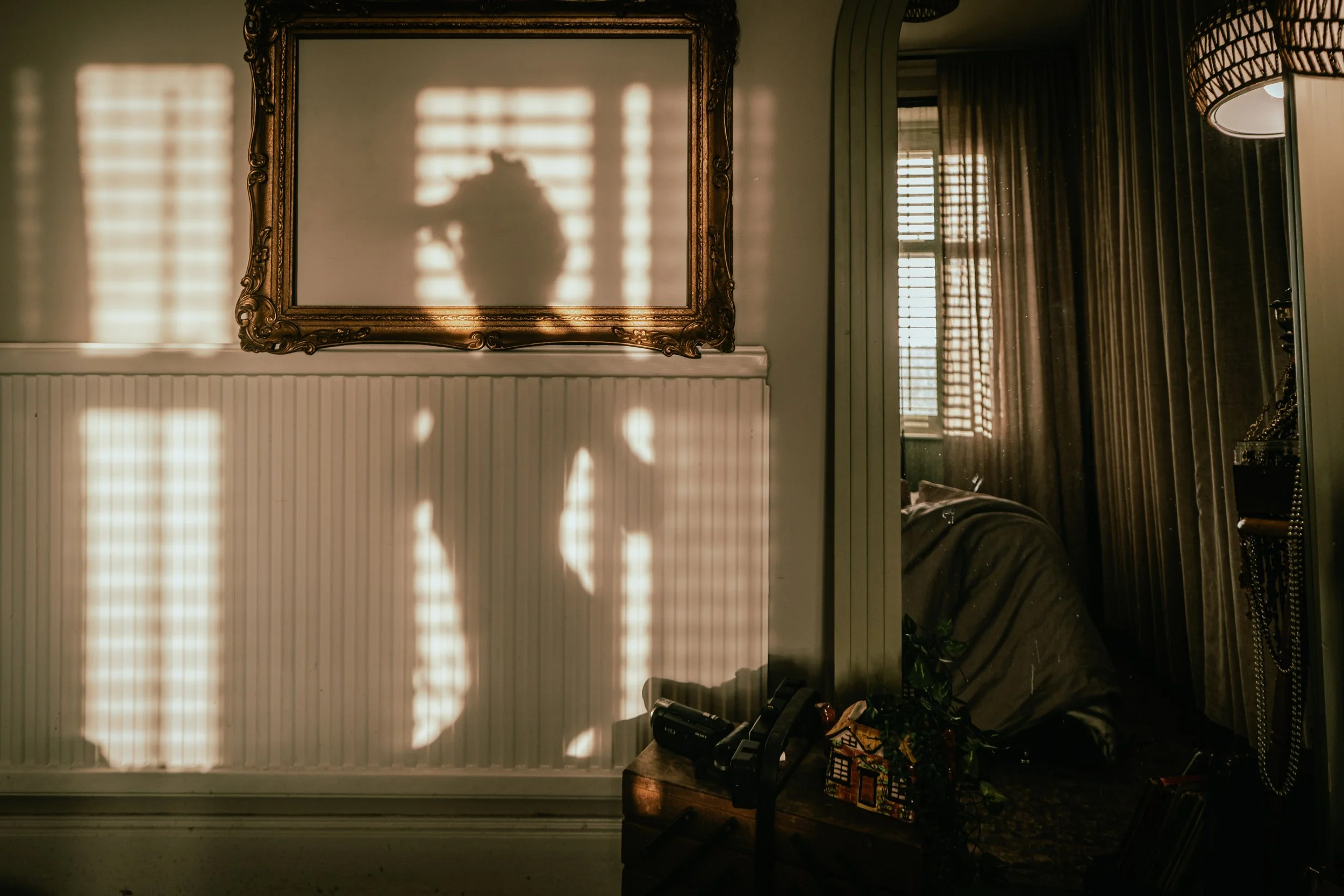 Sunlight casting shadows through window blinds onto a wall and mirror in a dimly lit room.