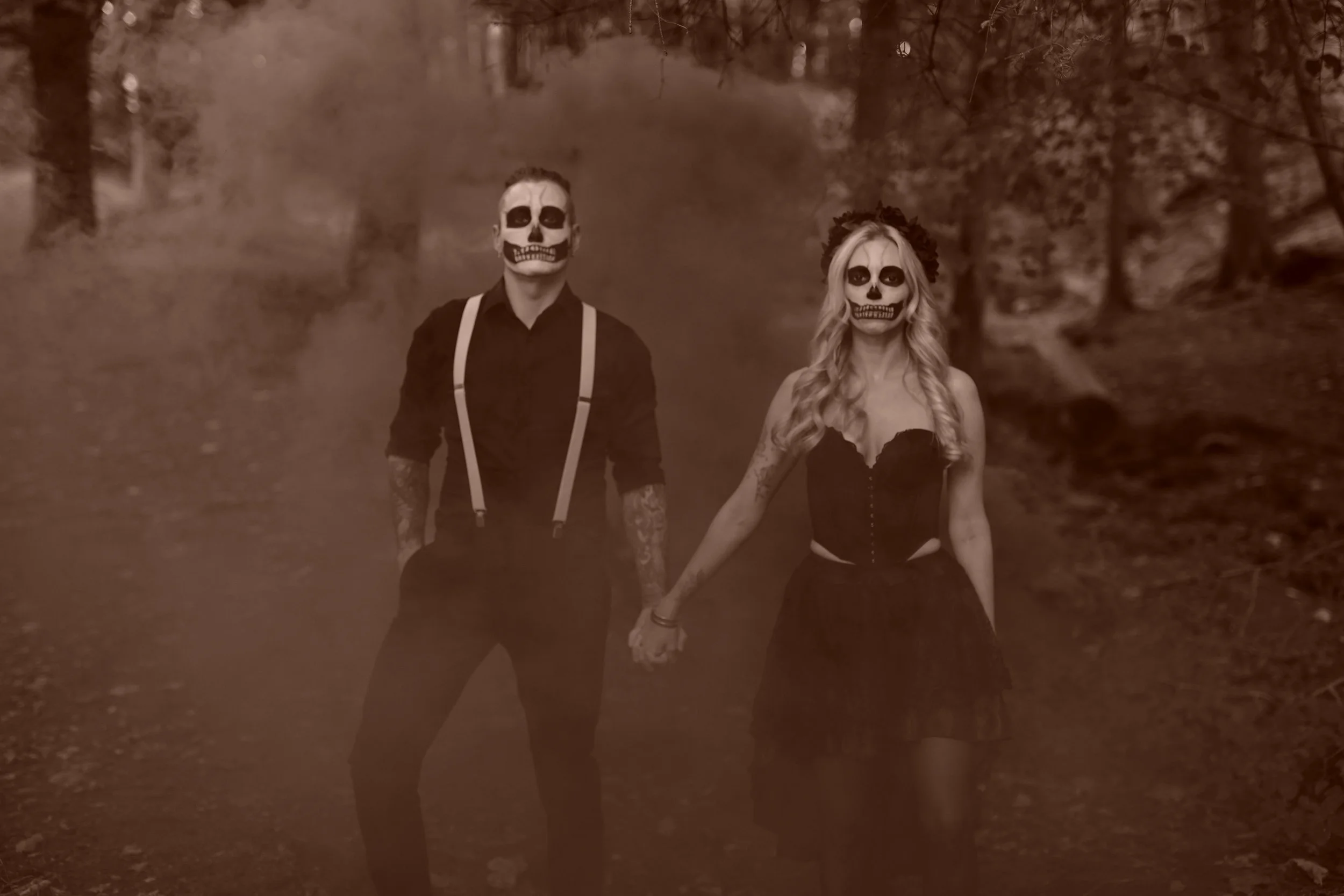 A man and woman with skeleton face paint holding hands standing outdoors at night in a wooded area.
