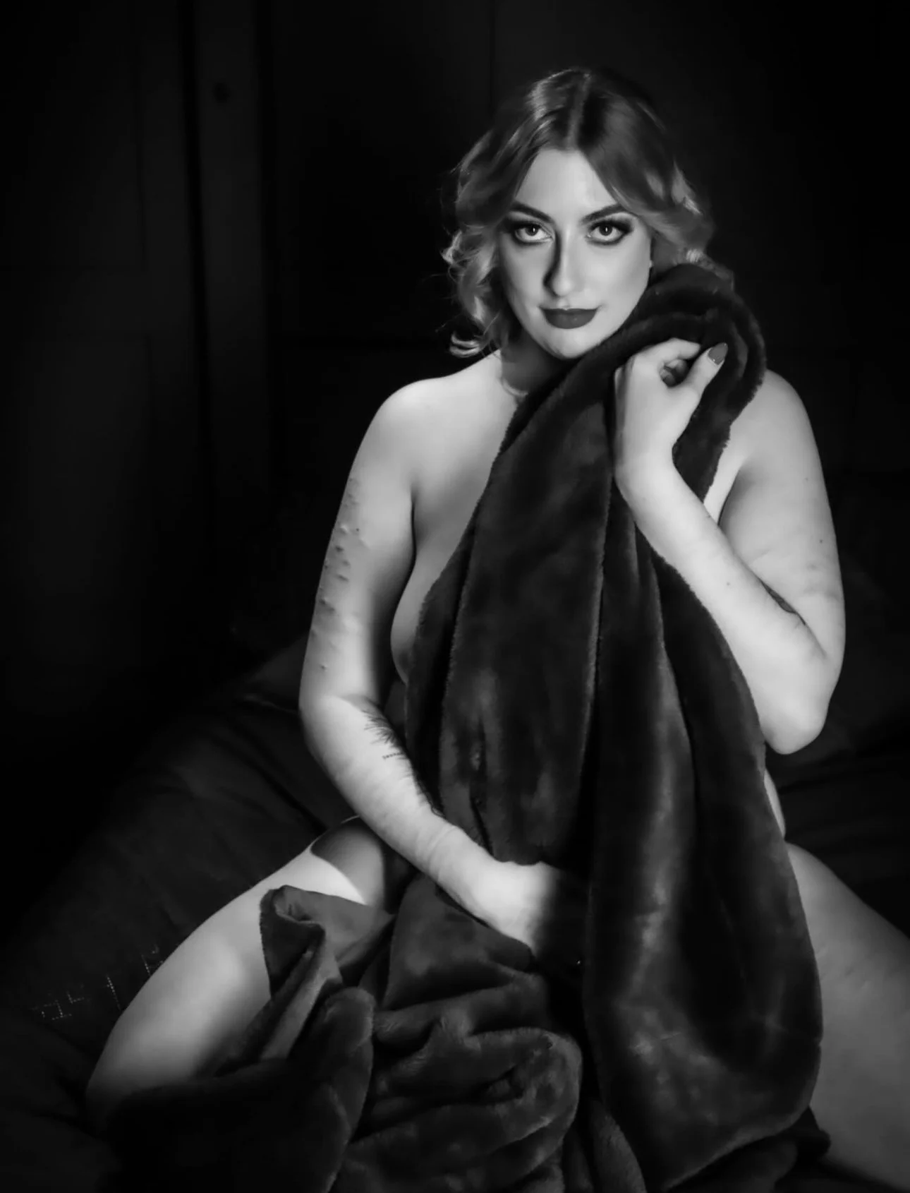 A woman with wavy hair and makeup poses topless, holding a fur coat across her chest in a dimly lit room.