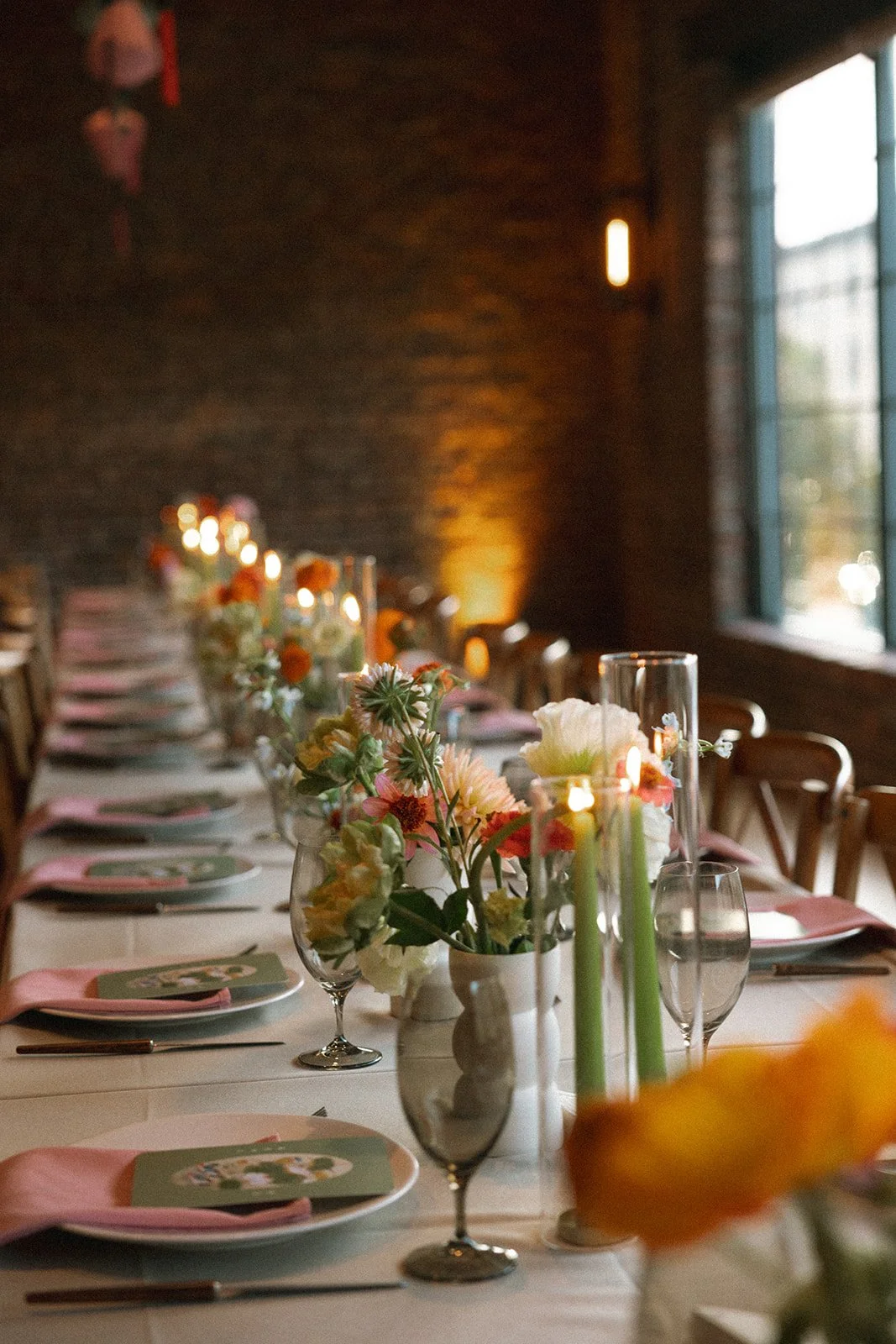 A long dining table set for a special occasion with floral arrangements, candles, wine glasses, and dinnerware in a warmly lit room with a brick wall and large window. marigolds florals nyc florist wedding florals