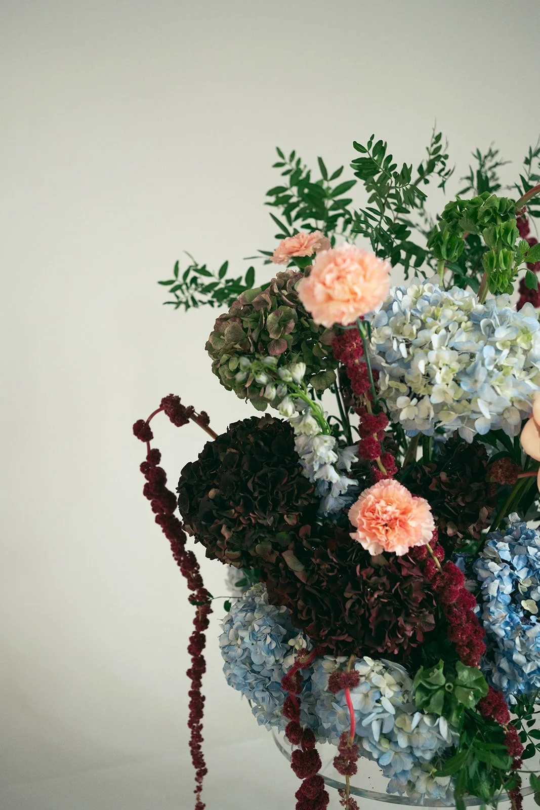 A close-up of a colorful bouquet of flowers, including pink carnations, light blue hydrangeas, dark red flowers, and green foliage, against a plain light background. marigolds florals nyc florist wedding florals atmosfera wedding