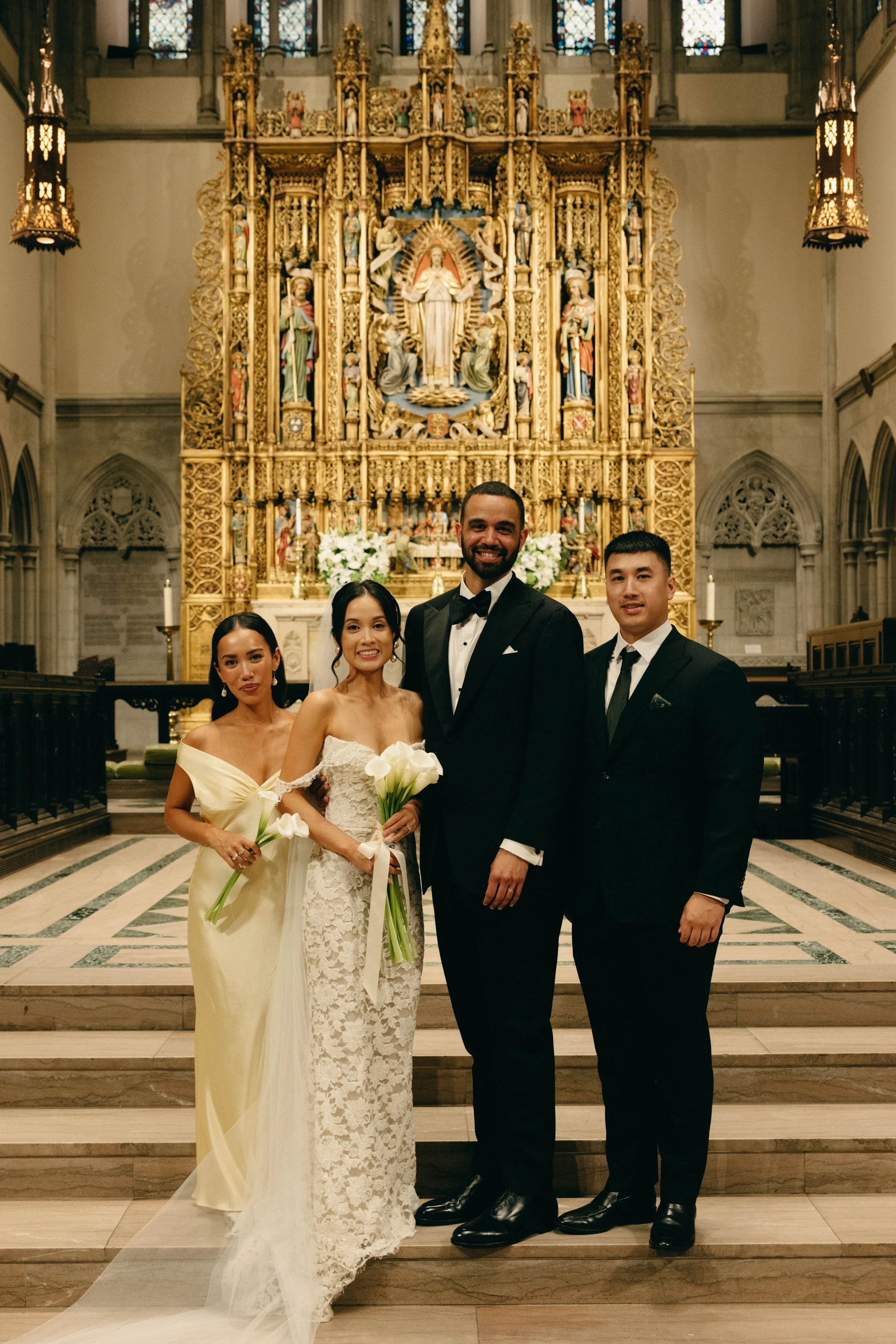 A wedding party standing in a church with a gold ornate altar in the background. The group includes two women and two men dressed in formal attire, holding white calla lilies, with the bride wearing a white lace wedding gown and the groom in a black 