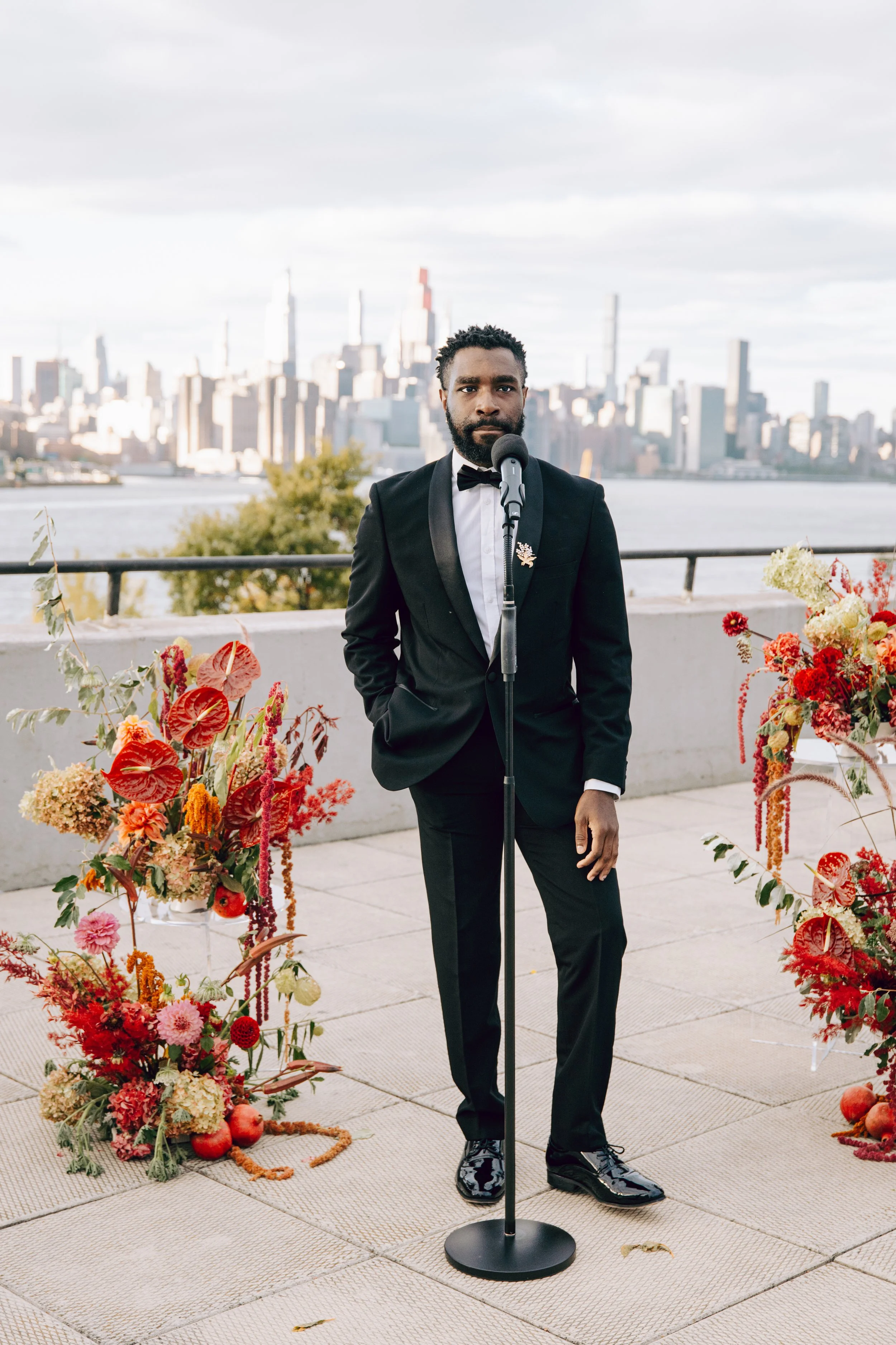 A man in a black tuxedo with a bow tie and a floral lapel pin stands in front of a microphone on a portable stand, outdoors near a city skyline, with colorful flower arrangements on either side. marigolds florals nyc florist wedding 