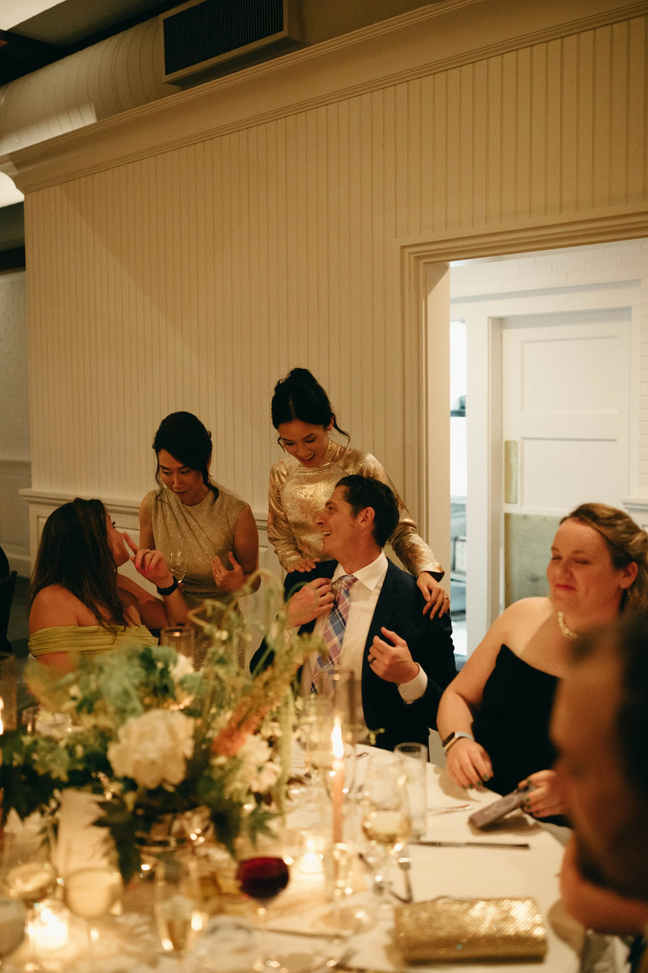Group of people at a formal dinner, with women in elegant dresses and a man in a tuxedo, seated at a table decorated with candles and floral arrangements. marigolds florals nyc florist wedding florals central park boathouse wedding