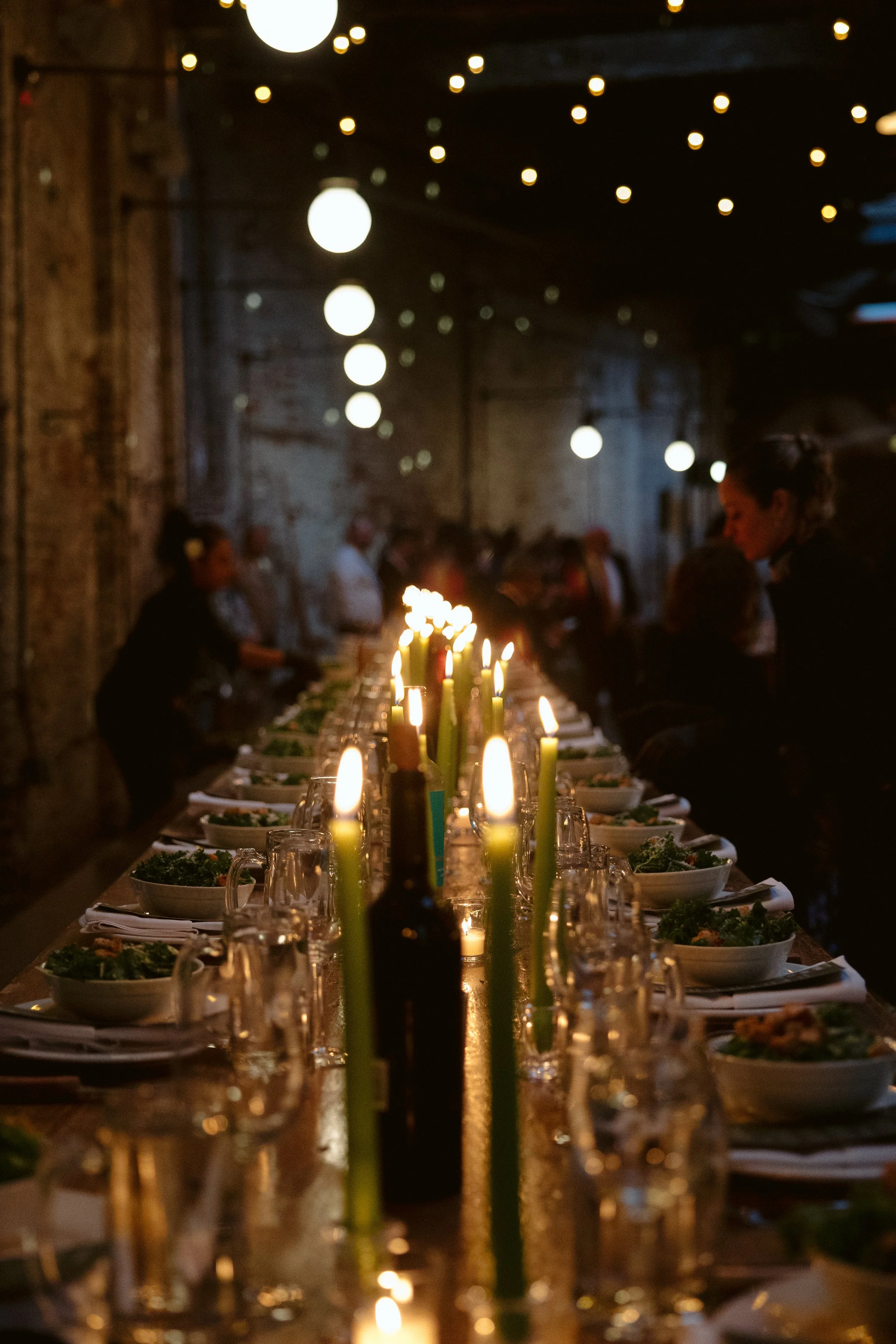 A long dining table set with plates of salad and glasses, illuminated by green candles and surrounded by people, in a dimly lit rustic setting with hanging string lights. marigolds florals nyc florist wedding florals houston hall nyc bar wedding