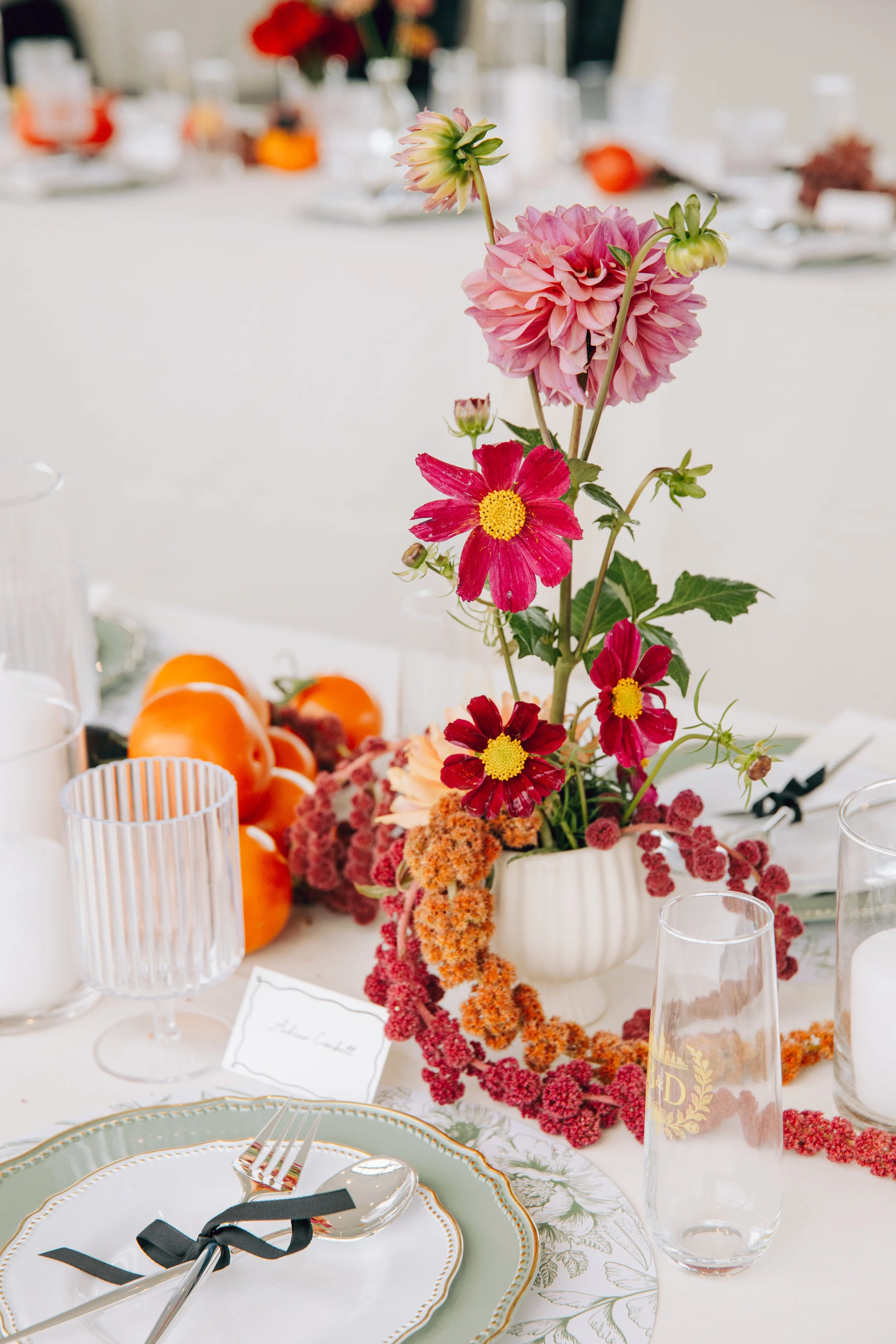 A floral centerpiece with pink and red flowers on a white table decorated with glassware, plates, and orange fruits. marigolds florals nyc florist wedding florals the w loft wedding pedestal wedding ceremony anthurium 