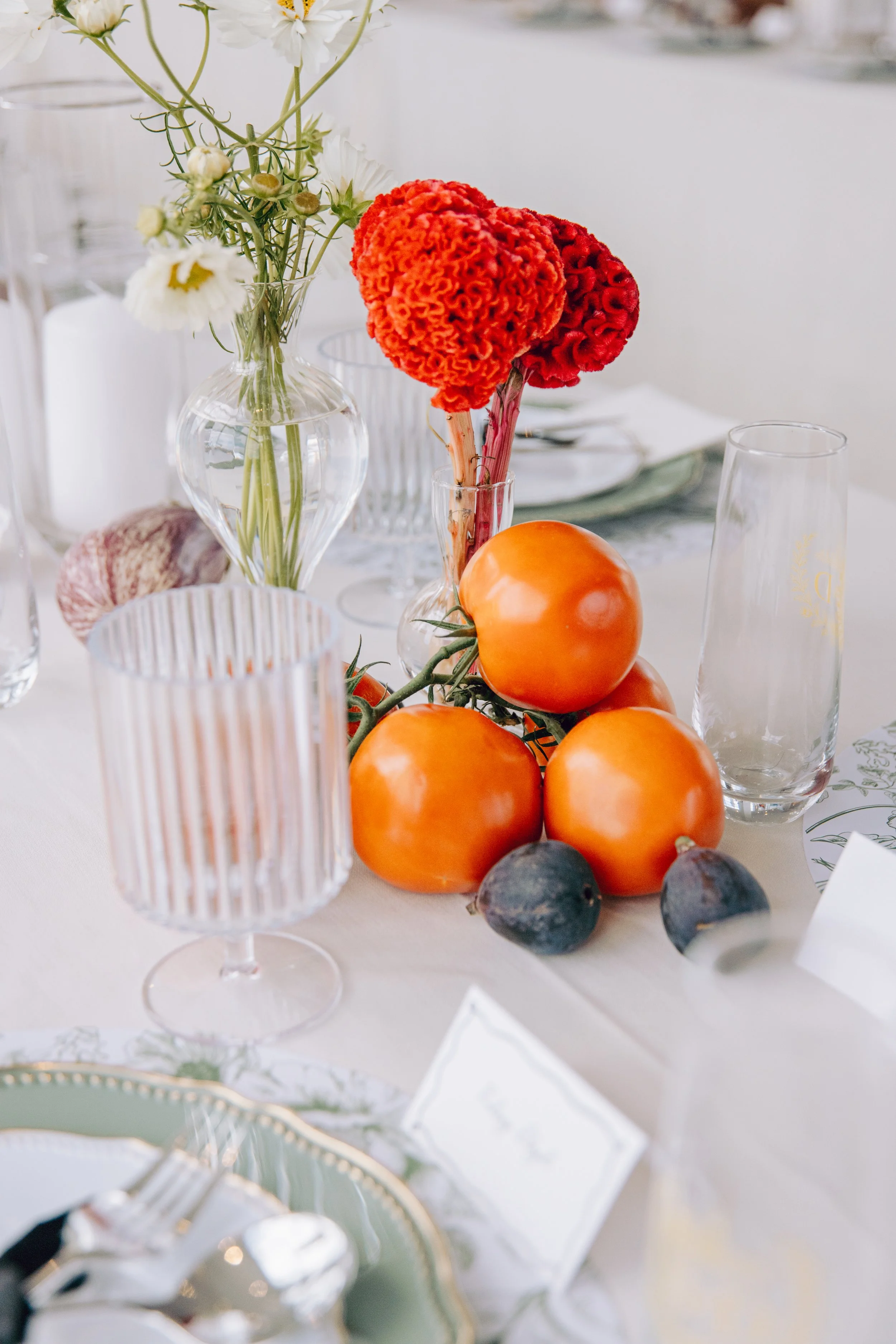 A table setting with a clear glass vase holding white flowers, surrounded by orange tomatoes and figs, with clear glasses and decorative plates nearby. marigolds florals nyc florist wedding florals the w loft wedding pedestal wedding ceremony