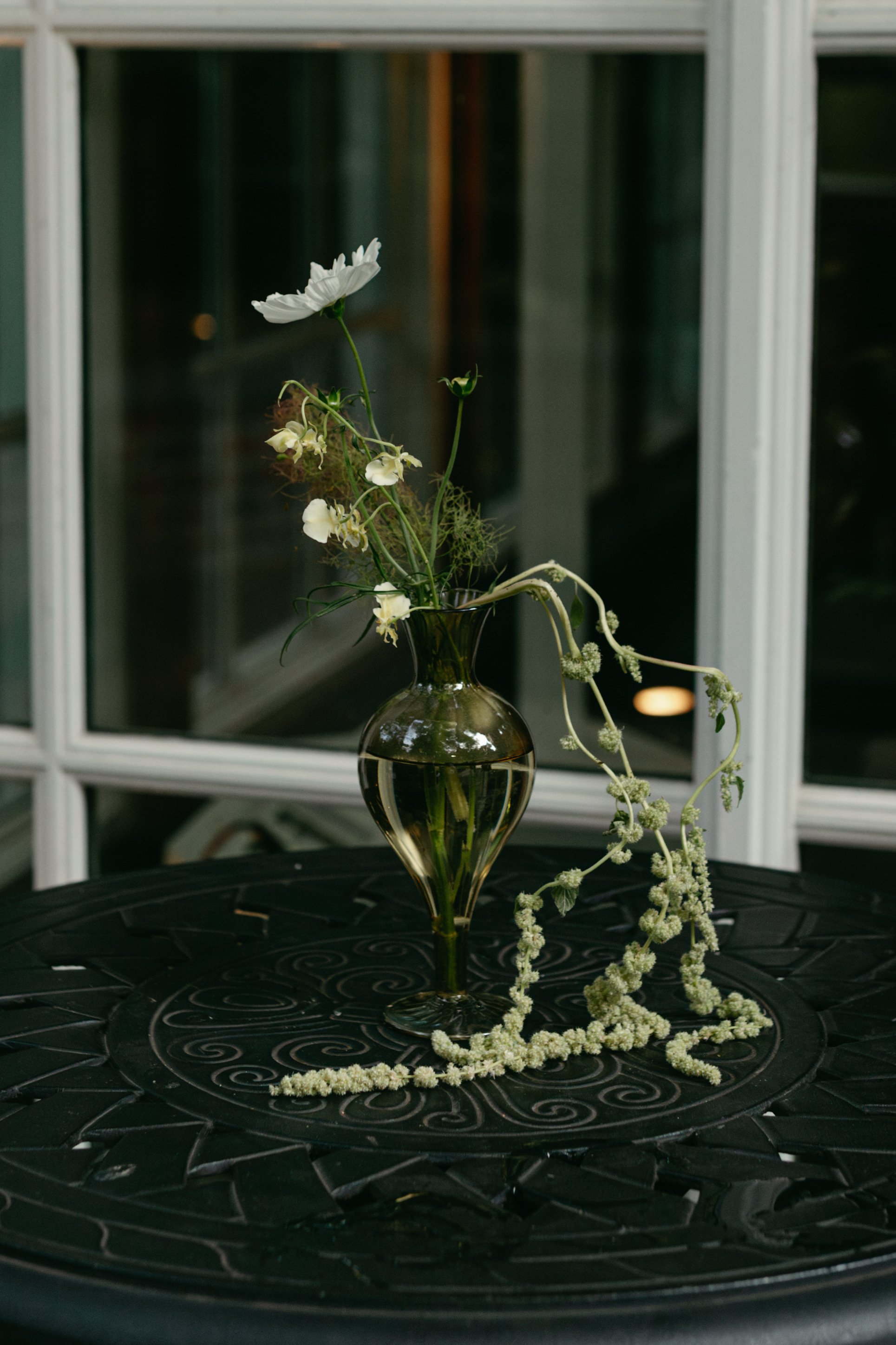 A dark glass vase with white flowers and trailing greenery on a black circular table with intricate swirl designs. In the background, there is a white-framed window with reflections of a dark interior. marigolds florals nyc florist wedding florals 