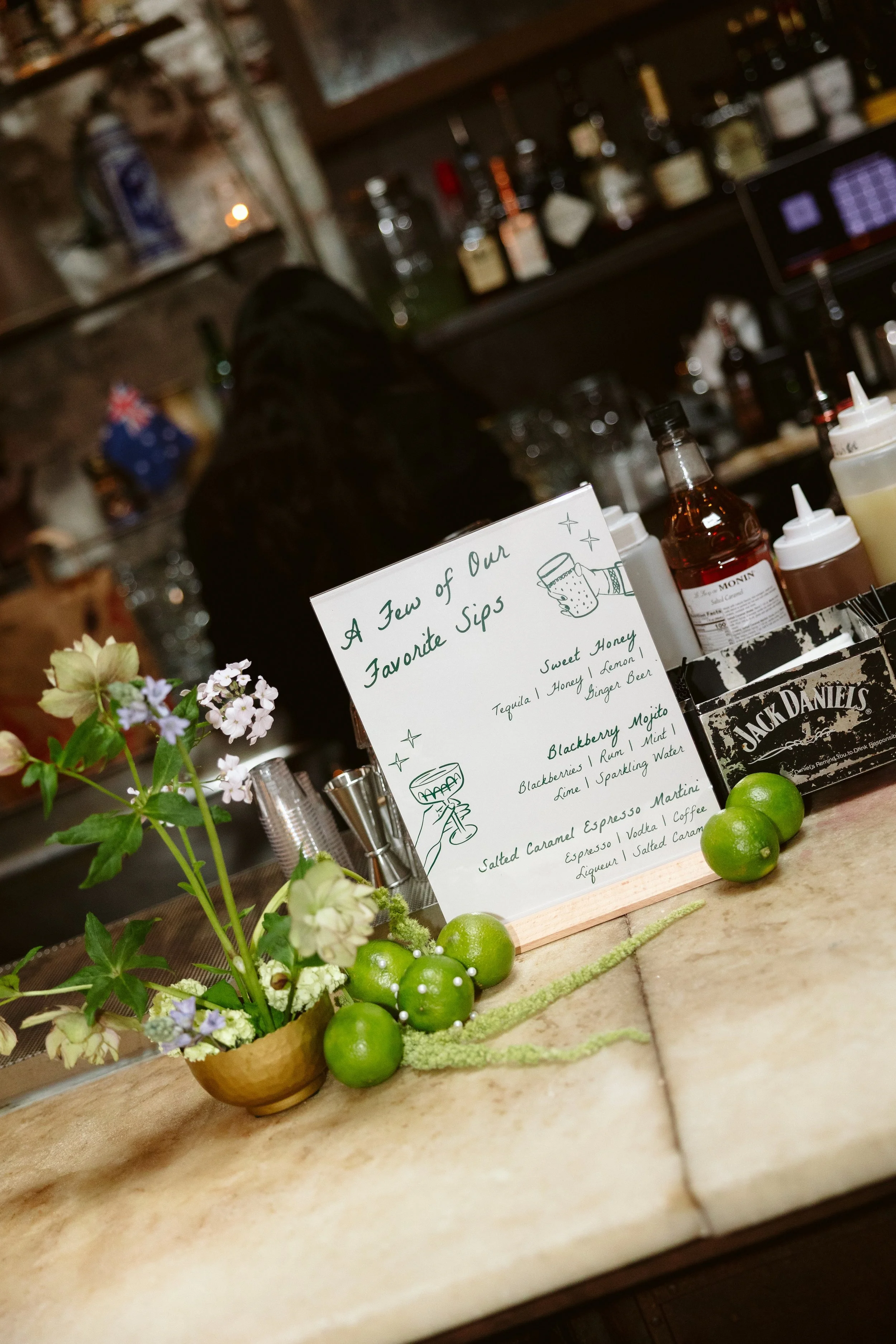 A bar counter with a handwritten drink menu, fresh flowers, and limes. The background has bottles of alcohol on shelves and a bartender behind the counter. marigolds florals nyc florist wedding florals houston hall nyc bar wedding