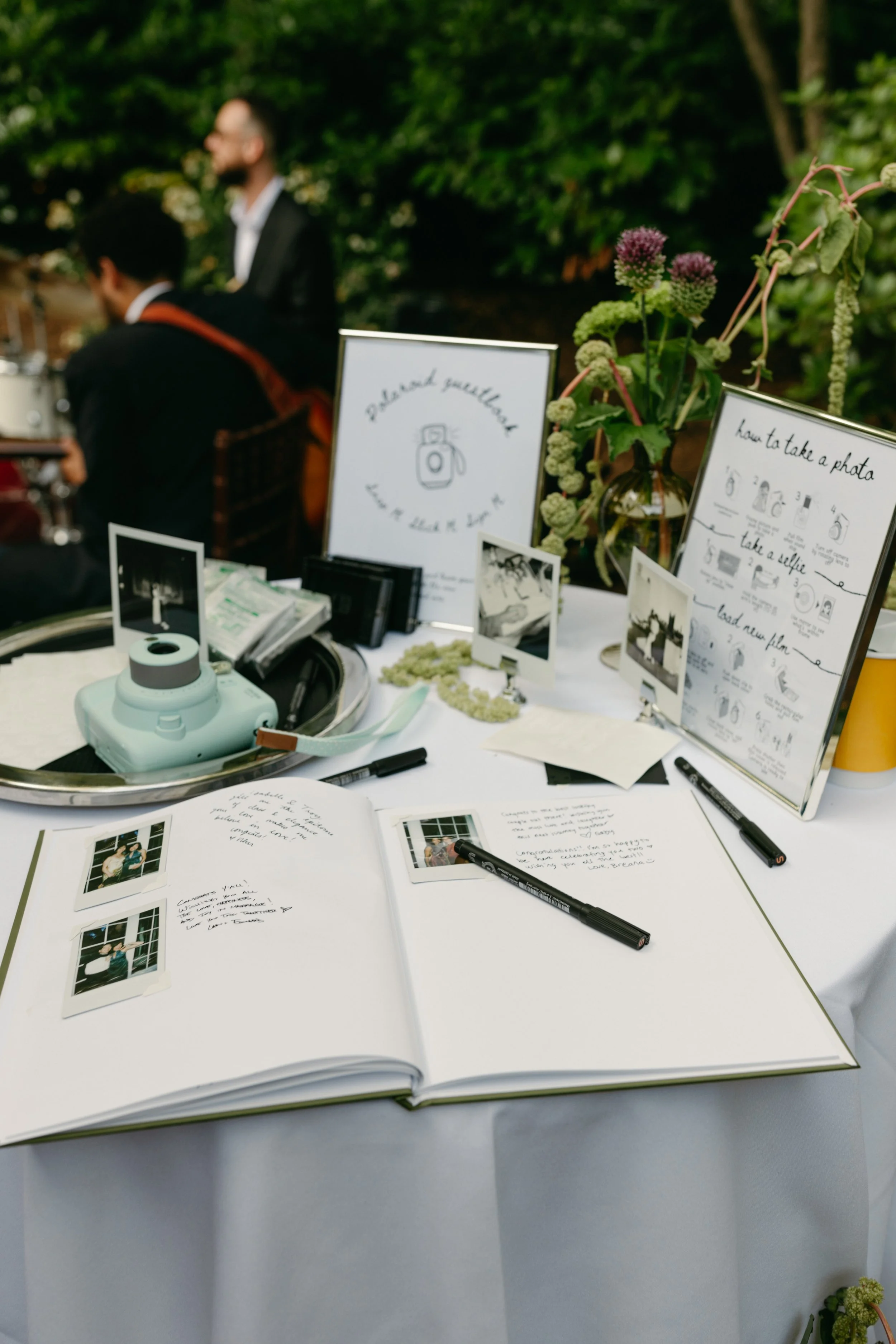 Open photo album on a white table with handwritten notes and Polaroid pictures, featuring two signs about taking and loading photos, and a floral arrangement in the background with two men in suits in an outdoor setting.  marigolds florals 