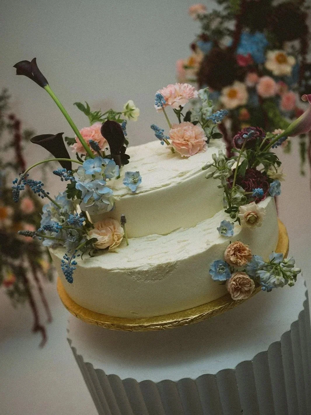 A two-tiered cake decorated with various fresh flowers including calla lilies, hydrangeas, and carnations, with floral arrangements on top and around the sides.