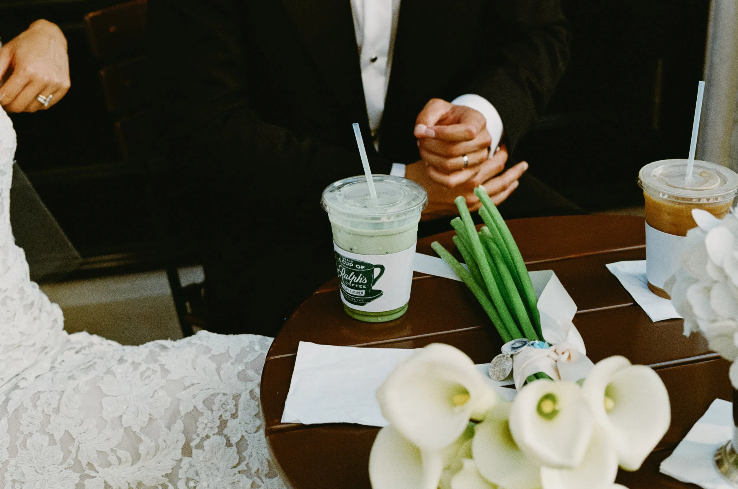 A close-up of a cake with white flowers in the foreground, with a partial view of a table with drinks and people seated around. Two clear plastic cups with green and caramel-colored drinks are visible on the table.