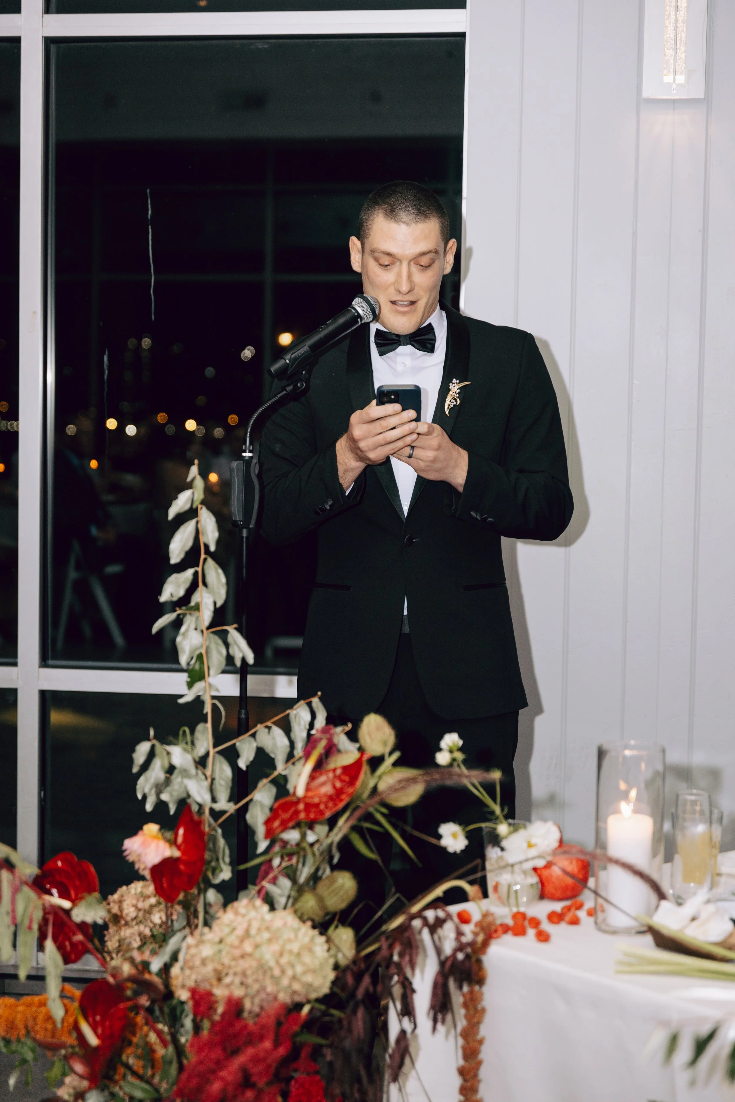 A man in formal attire with a bow tie is speaking or reading from a phone at a table decorated with flowers and candles during an indoor event. marigolds florals nyc florist wedding florals the w loft wedding pedestal wedding ceremony anthurium 