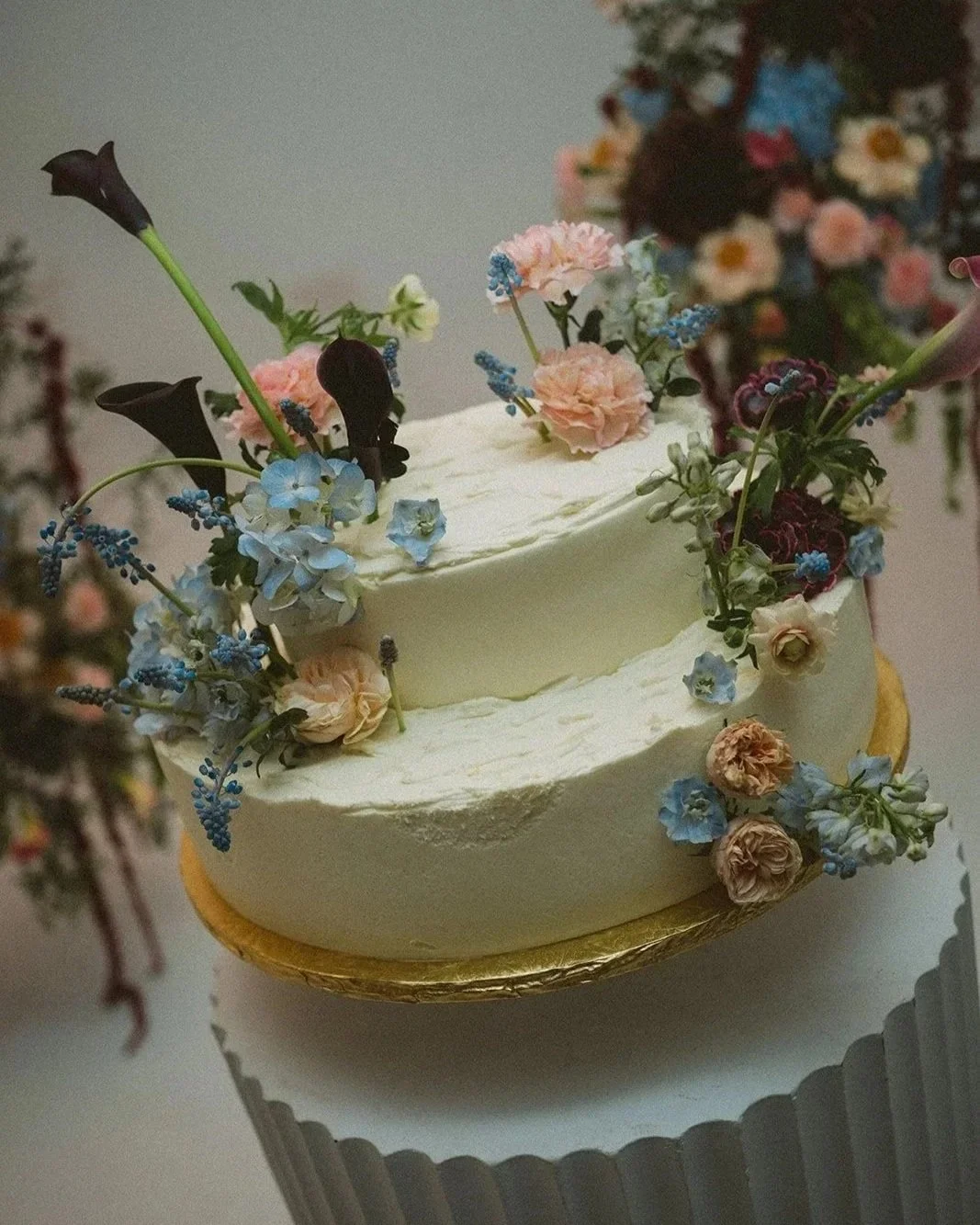 A white, two-tiered cake decorated with various pink, blue, and dark-colored flowers, on a gold cake board. Blues, pinks, purples wedding at Atmosfera in Queens New York. marigolds florals