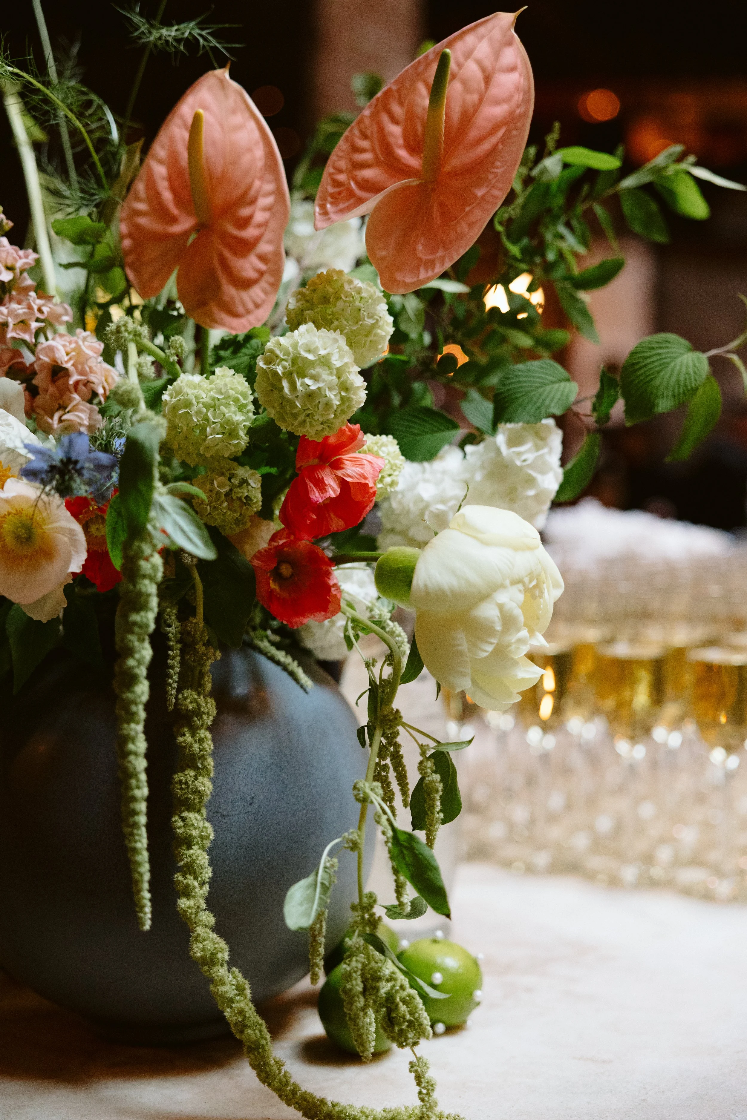 A floral arrangement with pink anthuriums, white hydrangeas, and green foliage in a dark gray vase on a wooden surface, with blurred lights in the background.