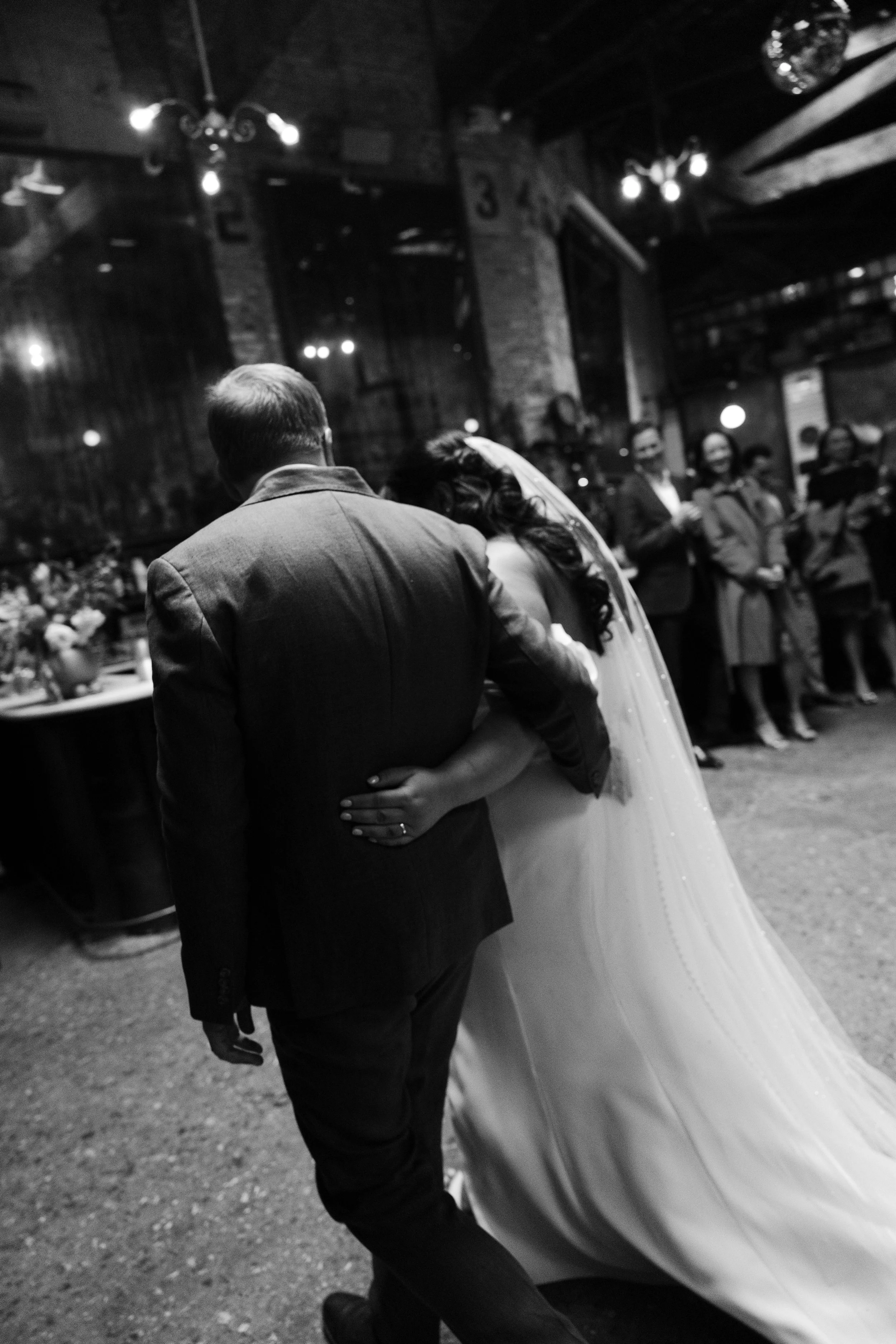 A black and white photo of a couple dancing close together inside a dimly lit venue, with several people in the background observing the dance. marigolds florals nyc florist wedding florals