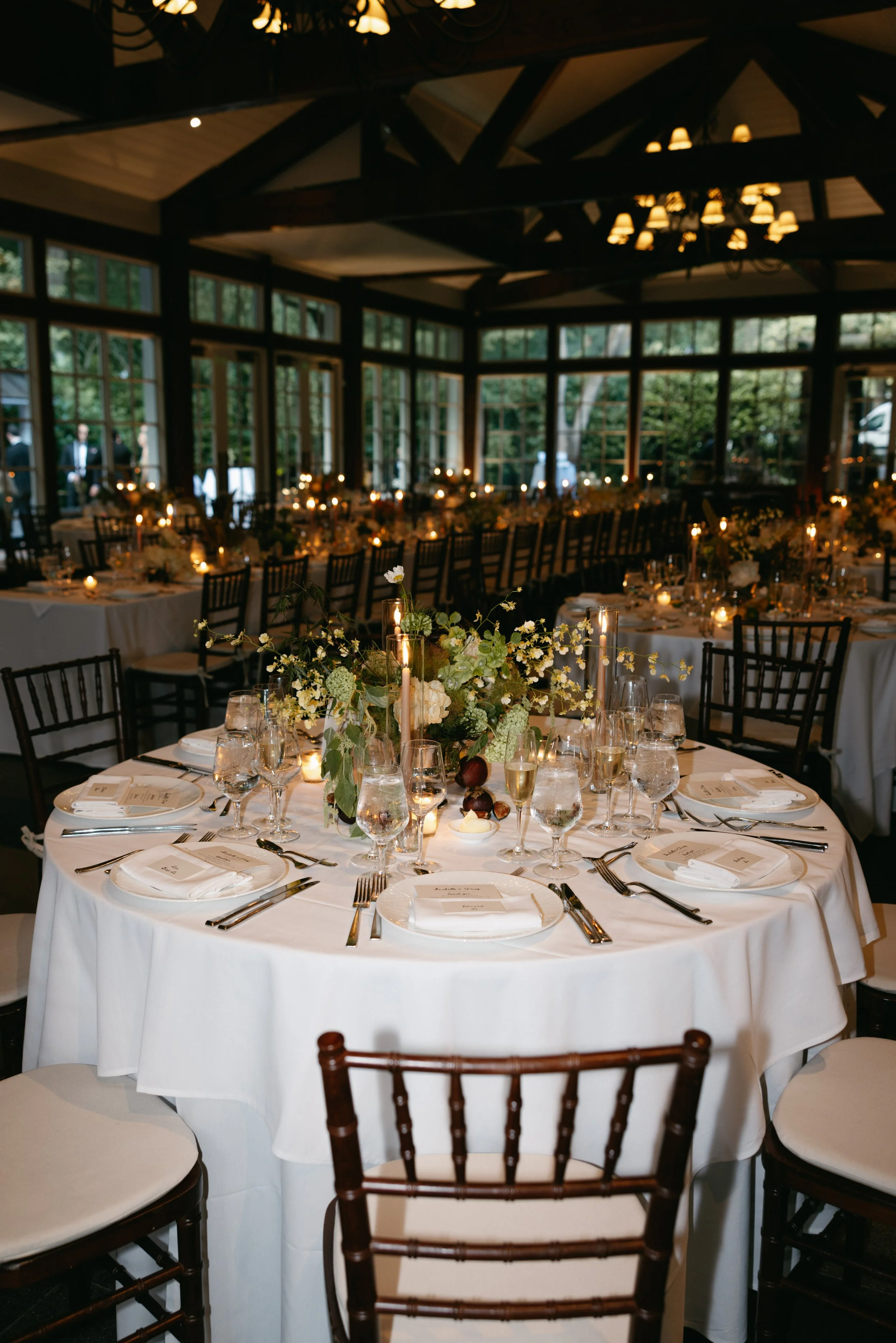 Elegant banquet table set for a formal event with white tablecloths, floral centerpieces, candles, glassware, and silverware, surrounded by dark wooden chairs in a glass-walled venue with green trees outside.