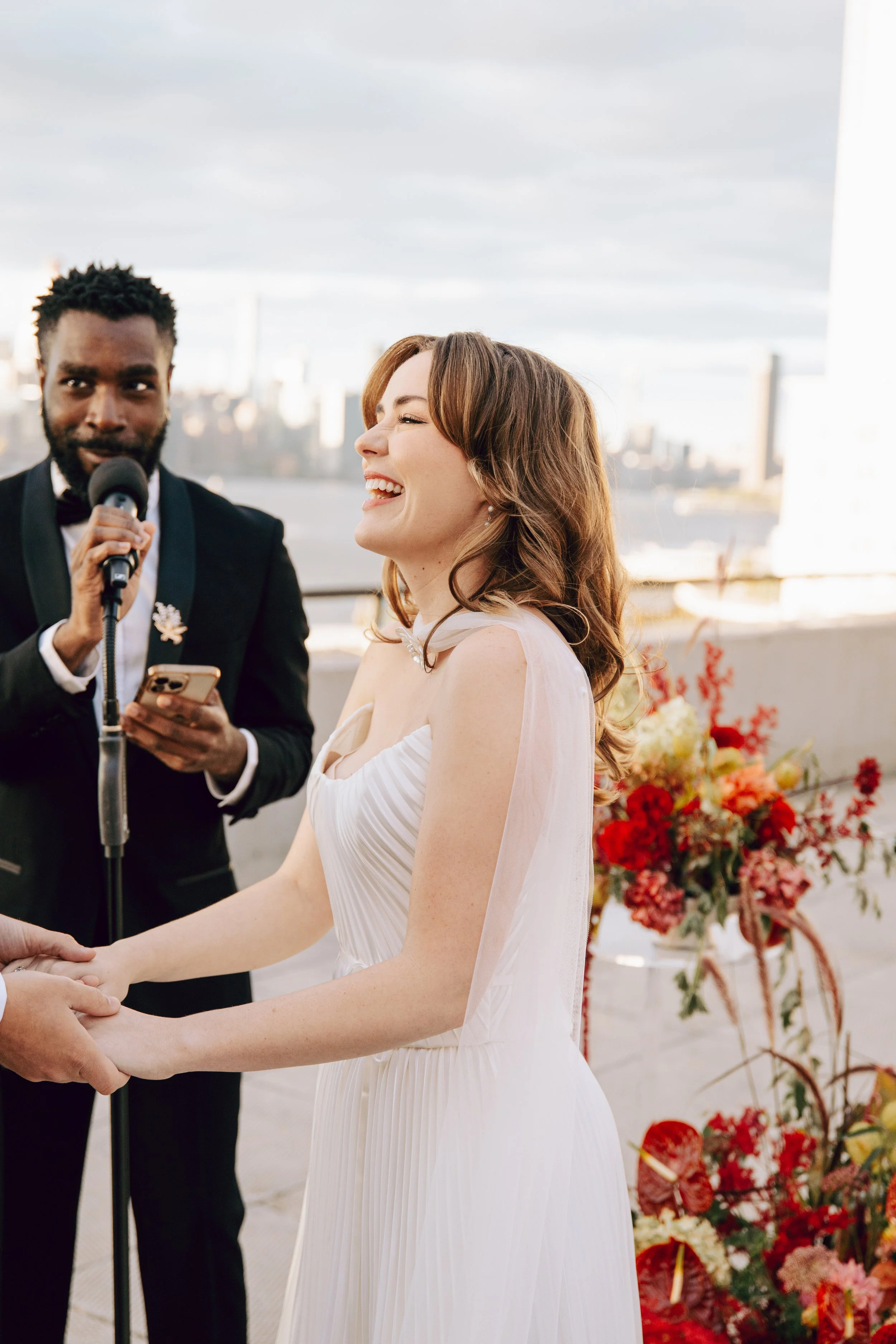 A woman in a white dress shares a joyful moment during a wedding ceremony, holding hands with someone off-camera. A man in a suit is speaking into a microphone nearby, possibly an officiant or best man. The background features a city skyline and a fl