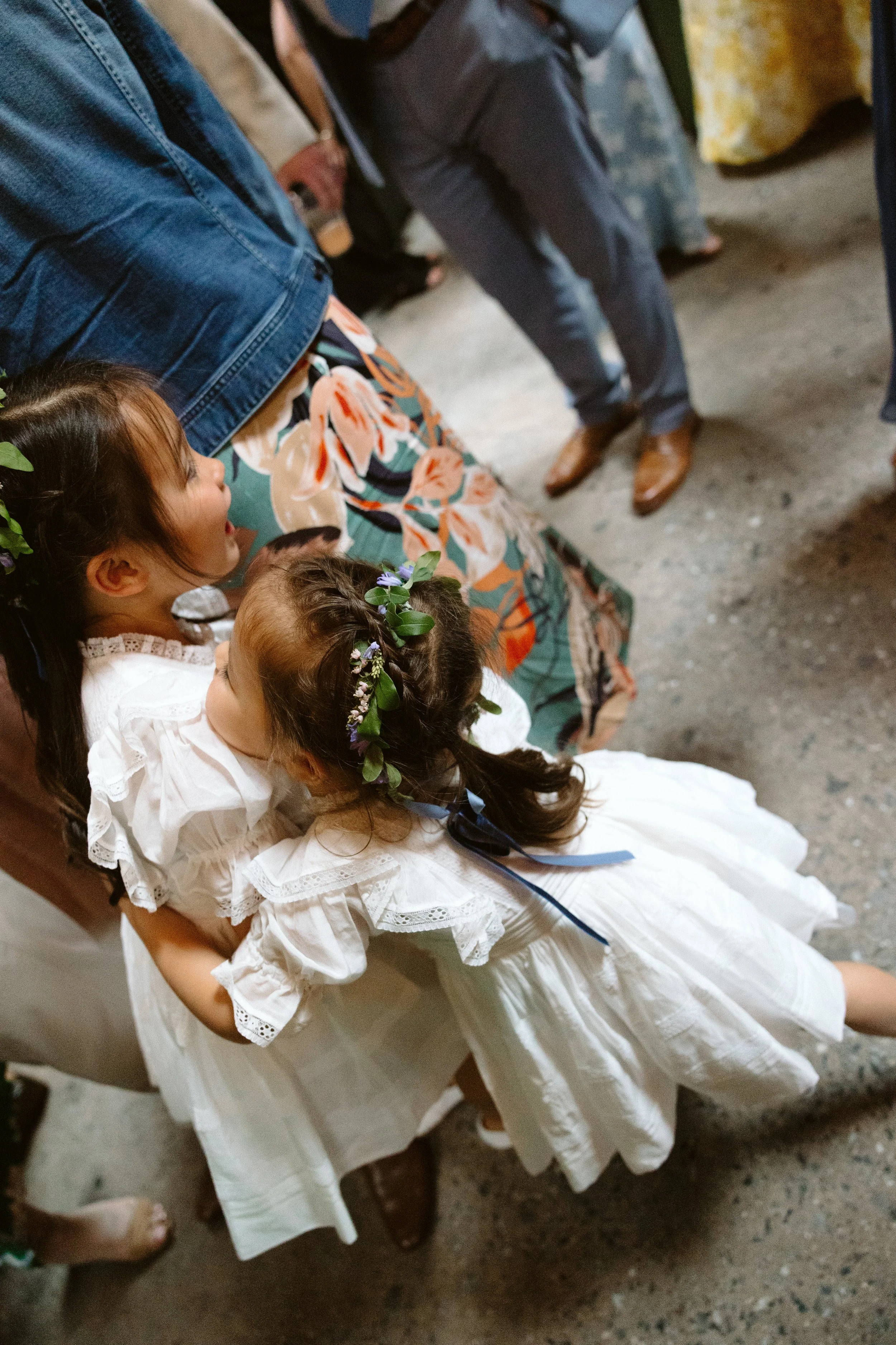 A girl in a white dress with lace details and floral crown on her head is sitting on a church pew, looking up at a man in formal attire. There are other people around her, some standing and some sitting, in the background.
