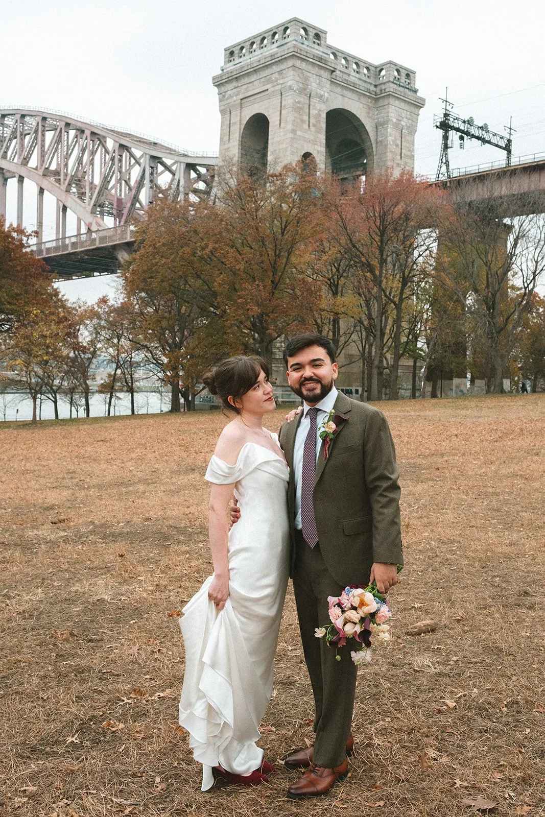 A bride and groom standing outdoors with autumn trees and a bridge in the background. marigolds florals nyc florist wedding florals atmosfera wedding