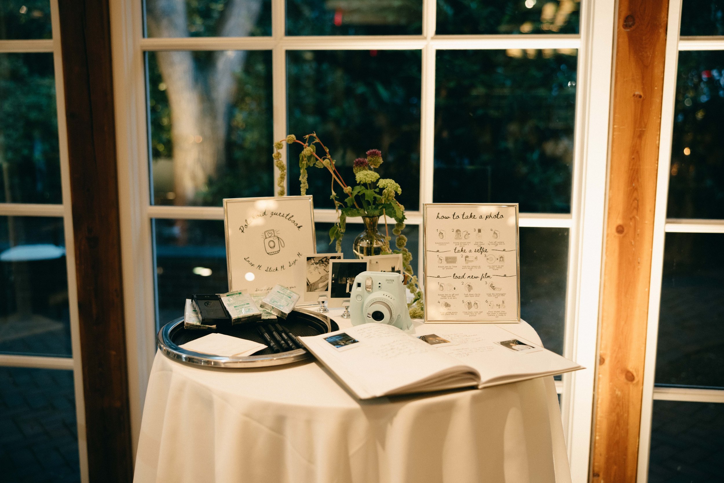 A table set up near a window with various items including a white instant camera, framed instructions on how to take photos, and a small vase with flowers. marigolds florals nyc florist wedding florals central park boathouse wedding