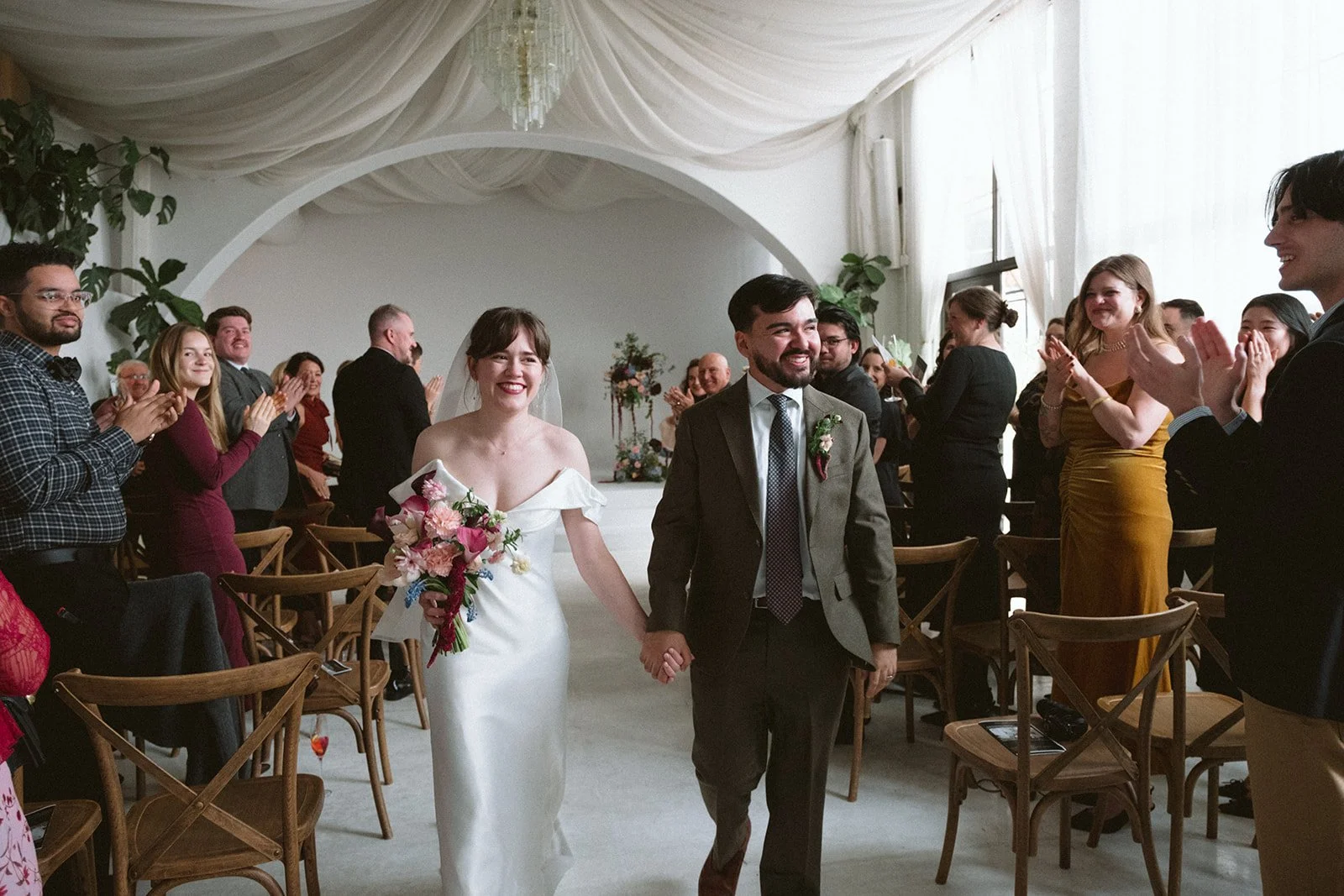 A bride and groom walk down the aisle holding hands, smiling, surrounded by cheering guests at a wedding ceremony in a decorated indoor venue. marigolds florals nyc florist wedding florals atmosfera wedding