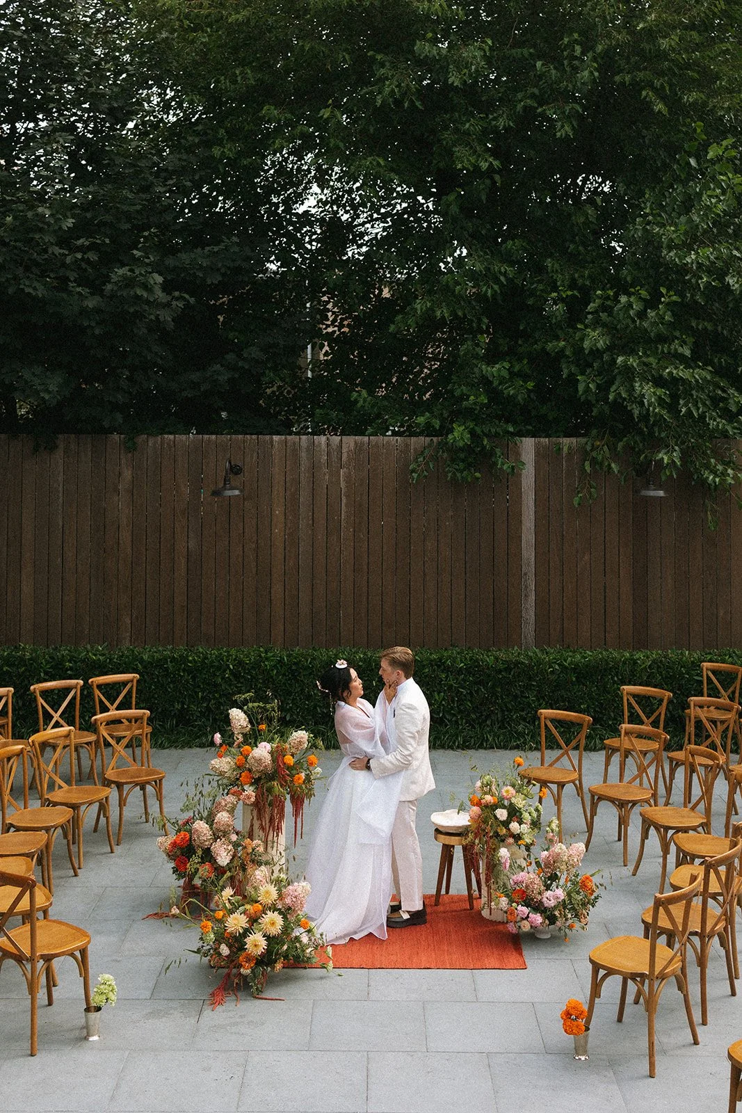 A bride and groom in wedding attire face each other on a small outdoor stage decorated with colorful flowers, surrounded by empty chairs, in a garden setting with tall trees and a wooden fence in the background. marigolds florals