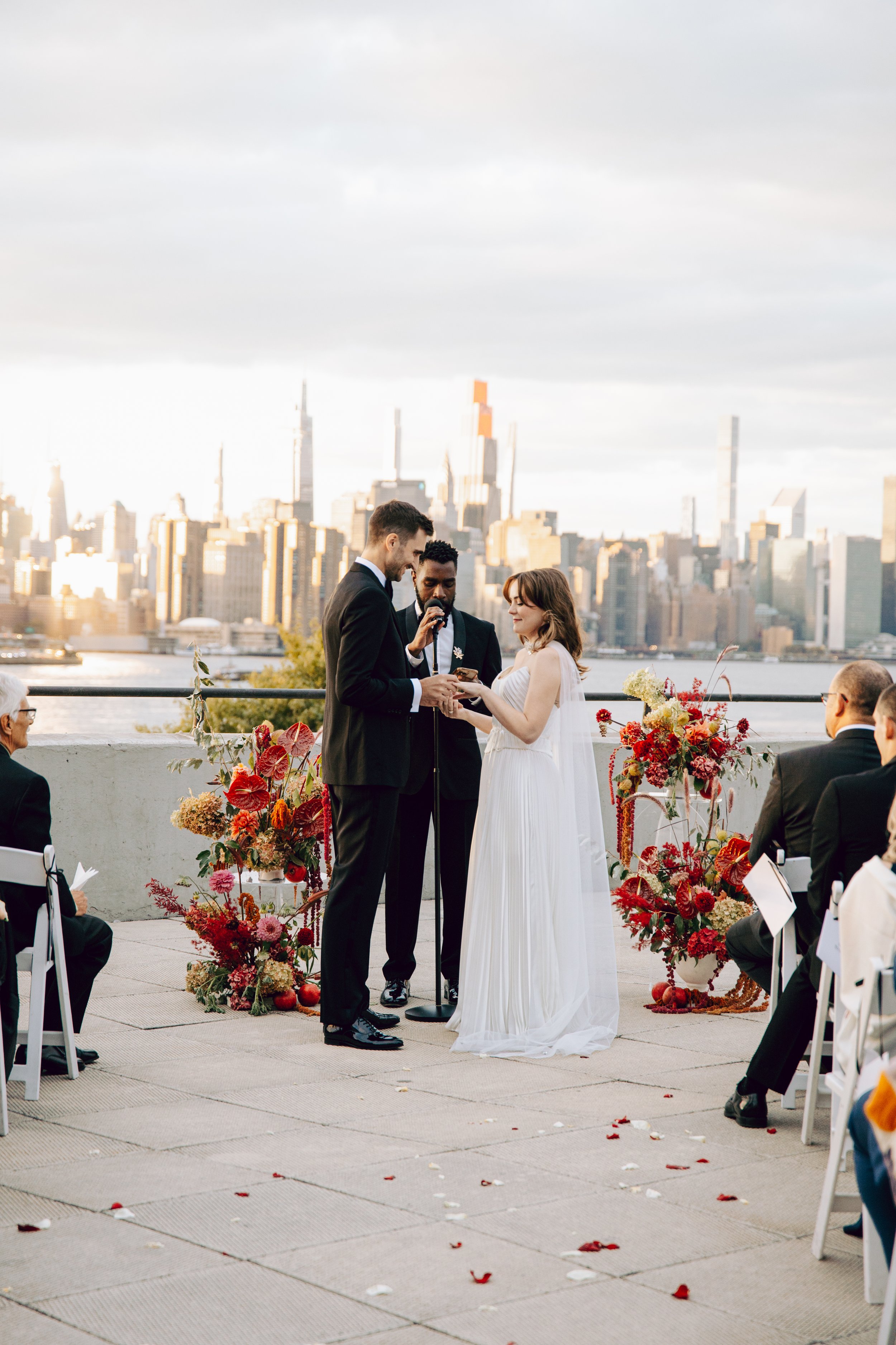 A wedding ceremony with a couple exchanging vows, surrounded by flowers, with a city skyline in the background during daylight. marigolds florals nyc florist wedding florals the w loft wedding pedestal wedding ceremony anthurium 