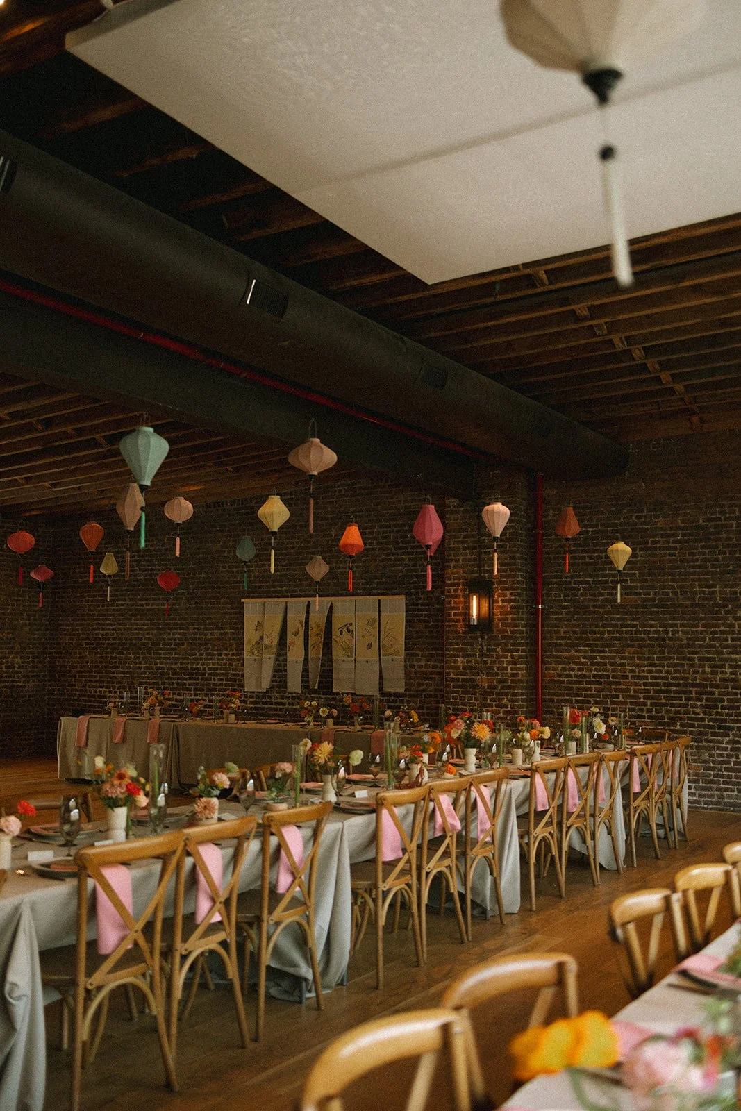 A decorated indoor event space with long dining tables, floral centerpieces, pastel napkins, and hanging paper lanterns, set against a brick wall with artwork. marigolds florals