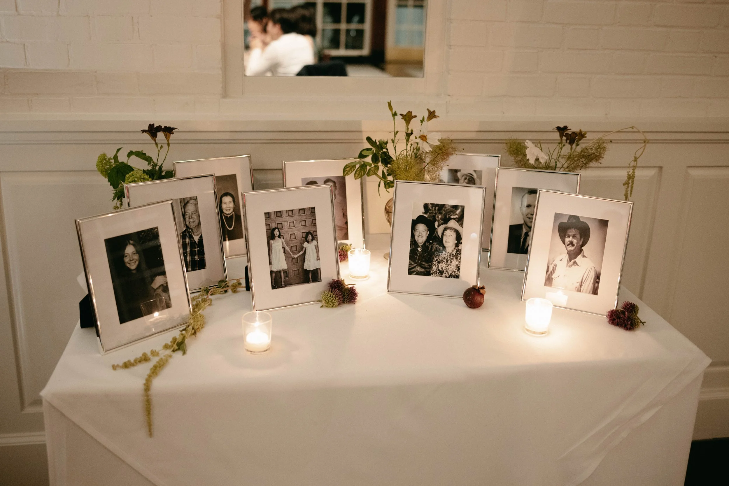 A memorial display with framed photographs of various people, some in color, some black and white, arranged on a white table. The display includes flowers and small candles, set against a white brick wall. A mirror above the display reflects people s