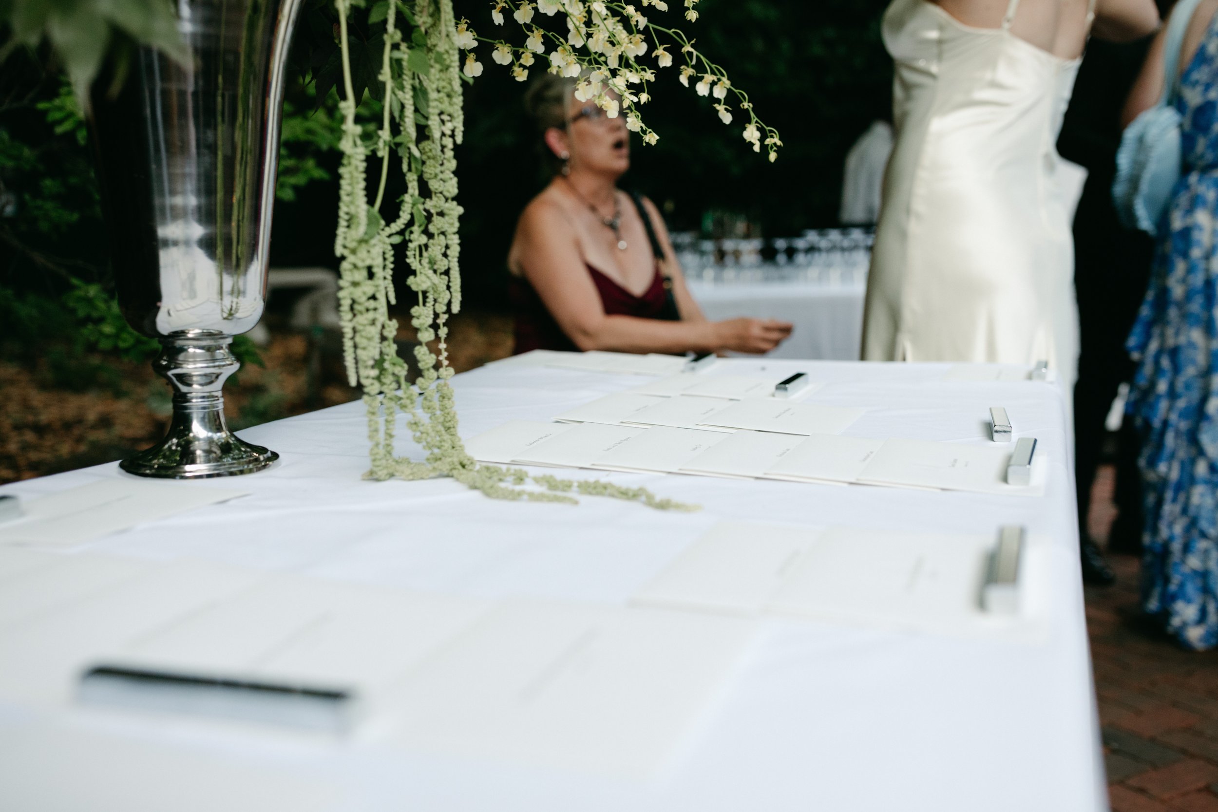 A white table with black and white printed papers and a large decorative silver vase with white hanging flowers, set outdoors with greenery in the background. A woman in a dark red dress is blurred in the background.