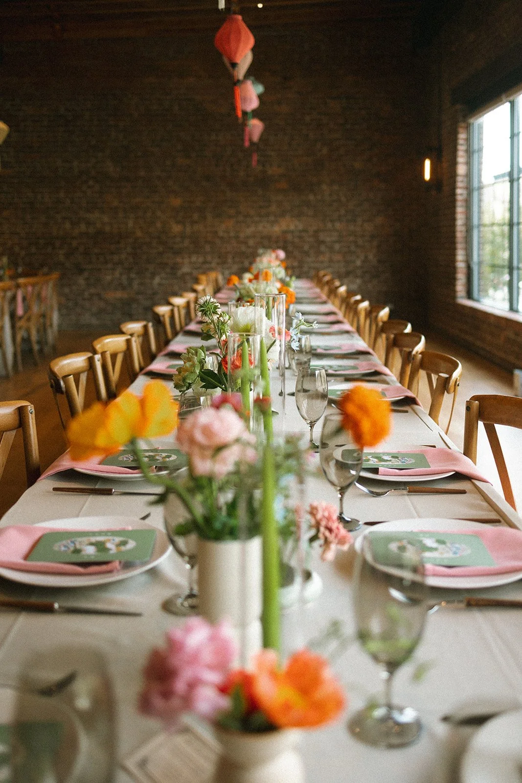 Long dining table set for an event with floral centerpieces, glasses, pink napkins, and place cards in a room with brick walls and large windows. marigolds florals nyc florist wedding florals