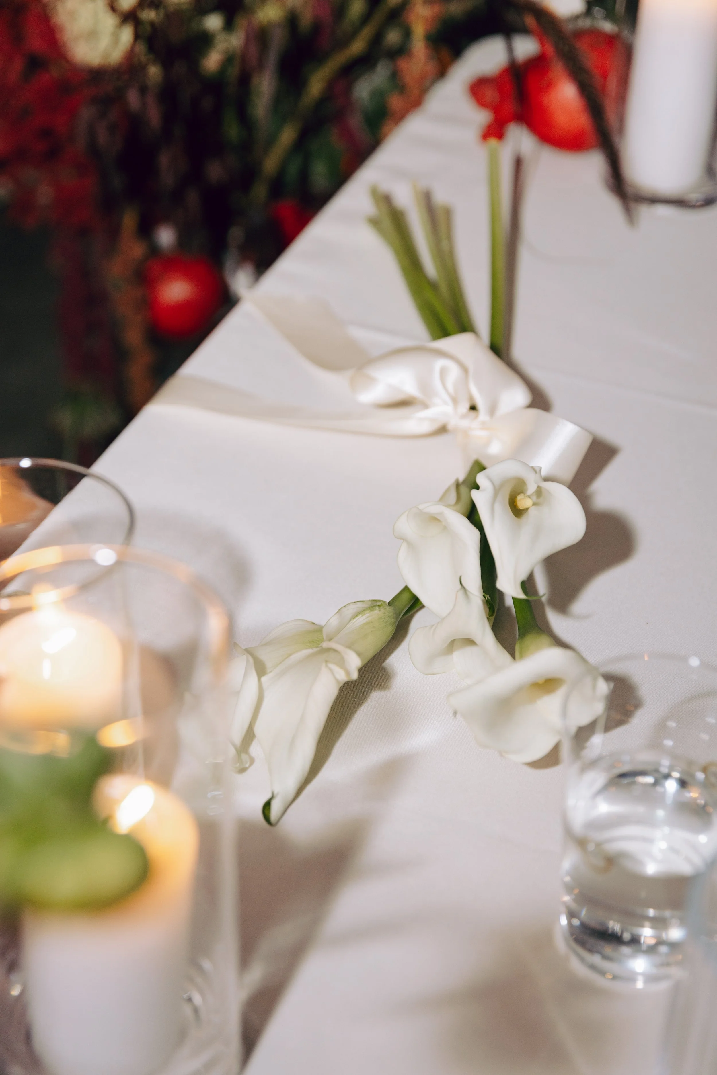 A white floral centerpiece with white calla lilies on a white tablecloth, surrounded by small glass candle holders with lit candles, against a blurred dark background with red and green colors.