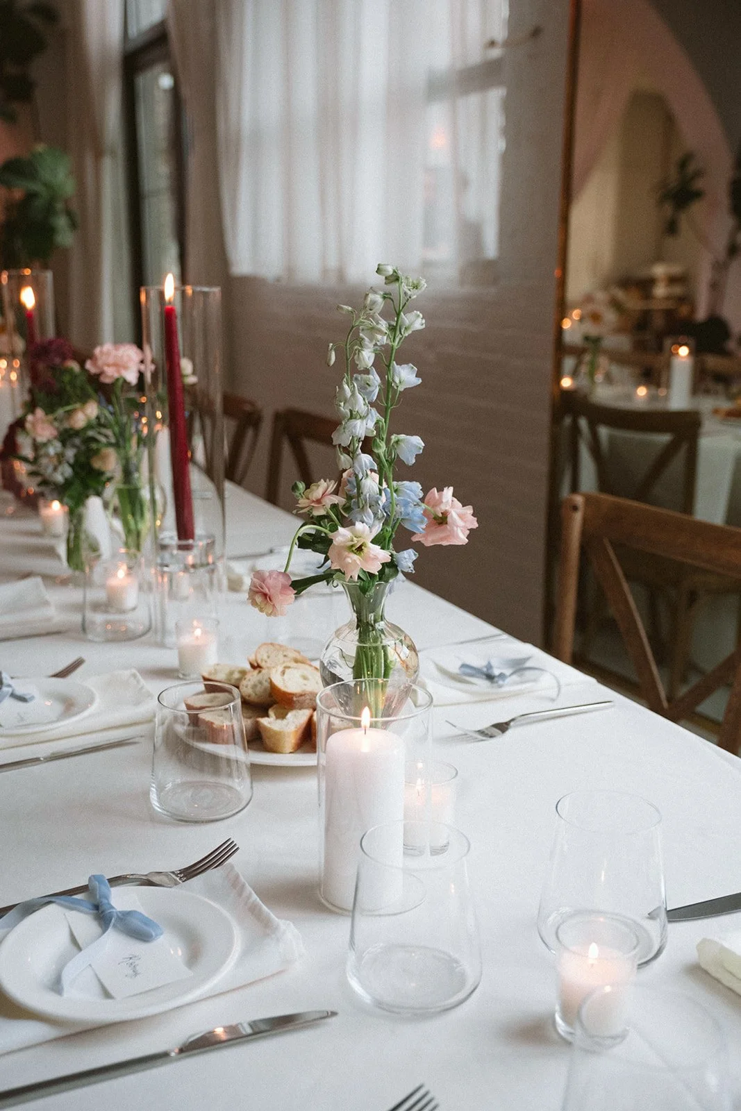 A dining table decorated with a white tablecloth, a floral centerpiece, candles, and place settings in a warmly lit room with curtains and wooden chairs. marigolds florals nyc florist wedding florals atmosfera wedding
