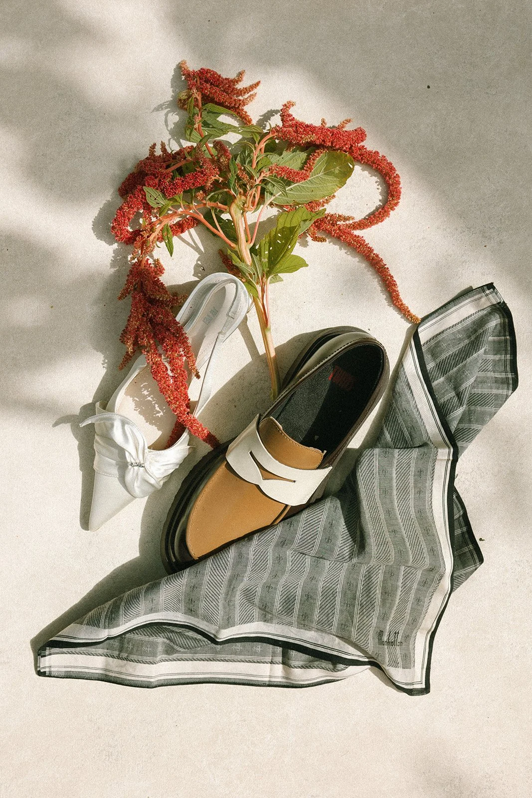 A white shoe, a brown and white loafer, and a checkered cloth with a branch of red flowers and green leaves placed on a light-colored surface. marigolds florals nyc florist wedding florals