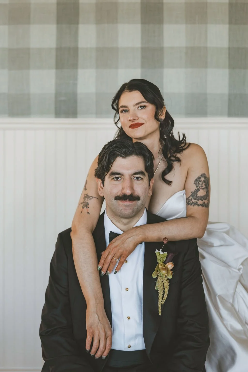 A bride and groom posing for a wedding photo, with the bride standing behind and resting her arms on the groom's shoulders, both smiling. The bride has dark hair, tattoos on her arms, and is wearing a white dress. The groom has dark hair, a mustache,
