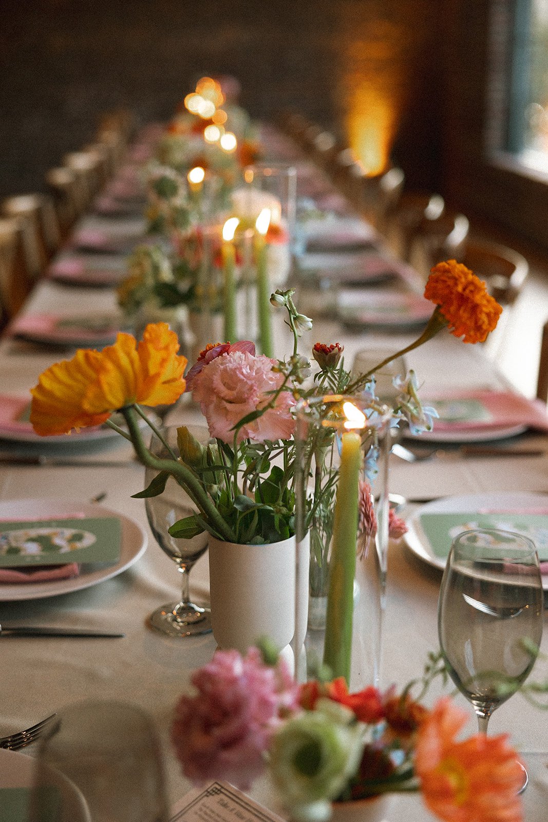 A long dining table decorated with colorful flowers in vases, lit candles, and place settings with pink napkins and printed menus, set in a warmly lit room. marigolds florals nyc florist wedding florals