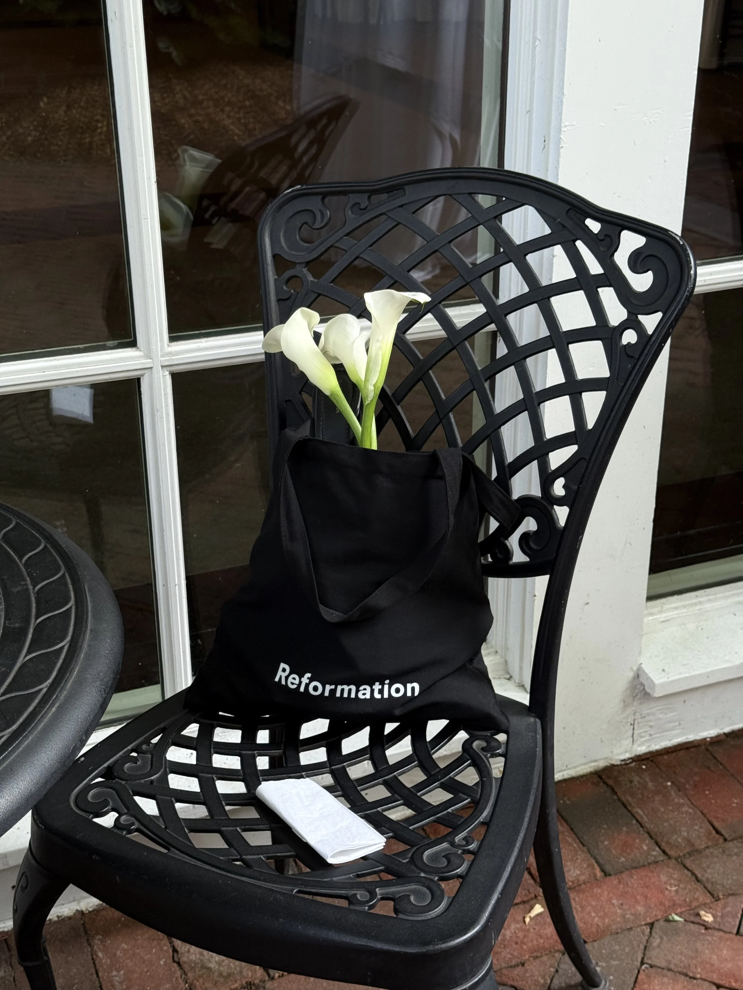 Black metal outdoor chair with white calla lilies and a Reformation cloth bag on the seat, situated in front of a glass door with white framing on a brick patio. marigolds florals nyc florist wedding florals central park boathouse wedding